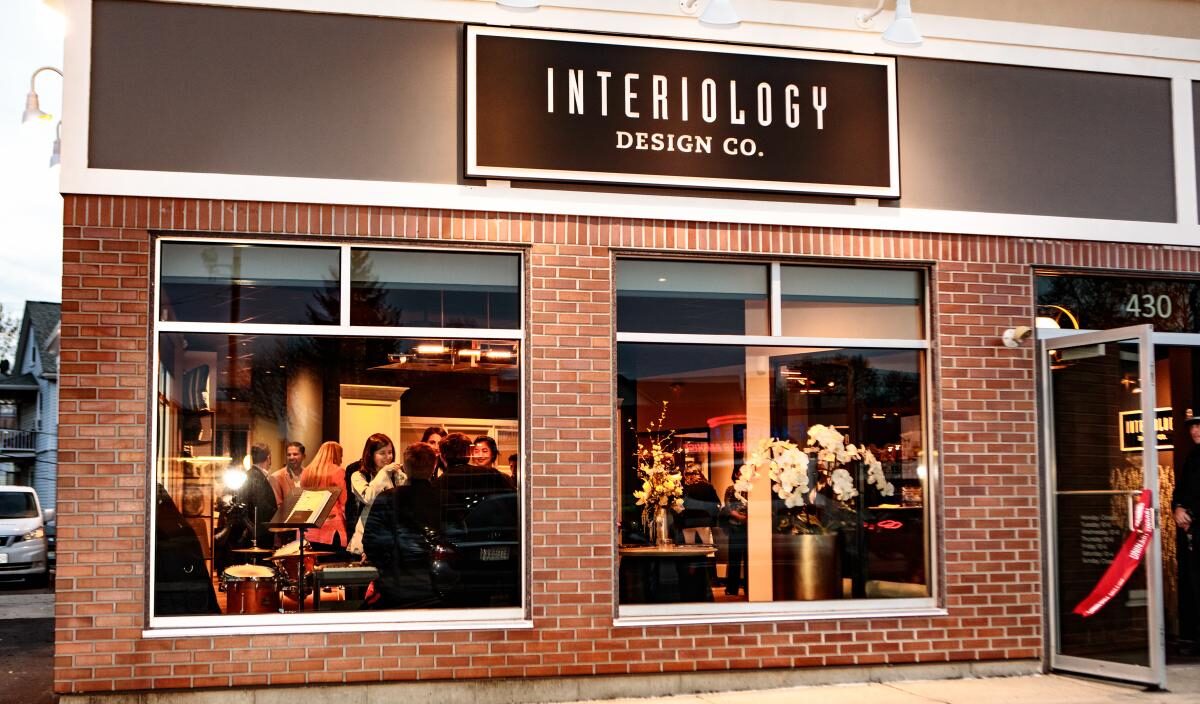 People gather inside Interiology Design Co., visible through large windows of a brick storefront with the company sign above and floral decorations near the entrance, offering a glimpse into what’s special about Interiology.