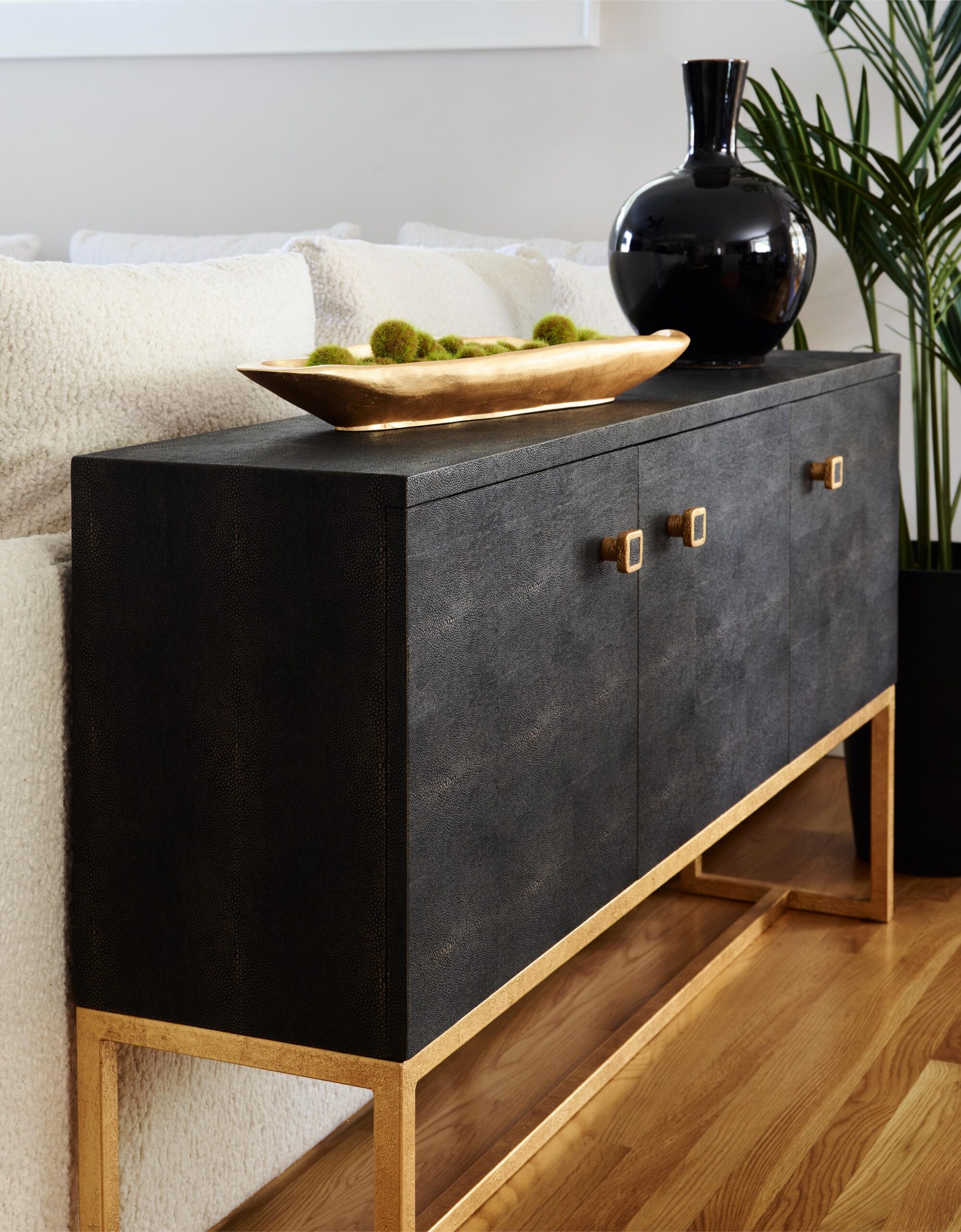 A black sideboard with gold trim and square handles stands on a wooden floor in the living space, topped with a gold bowl, green decorative balls, and a black vase next to a leafy plant.