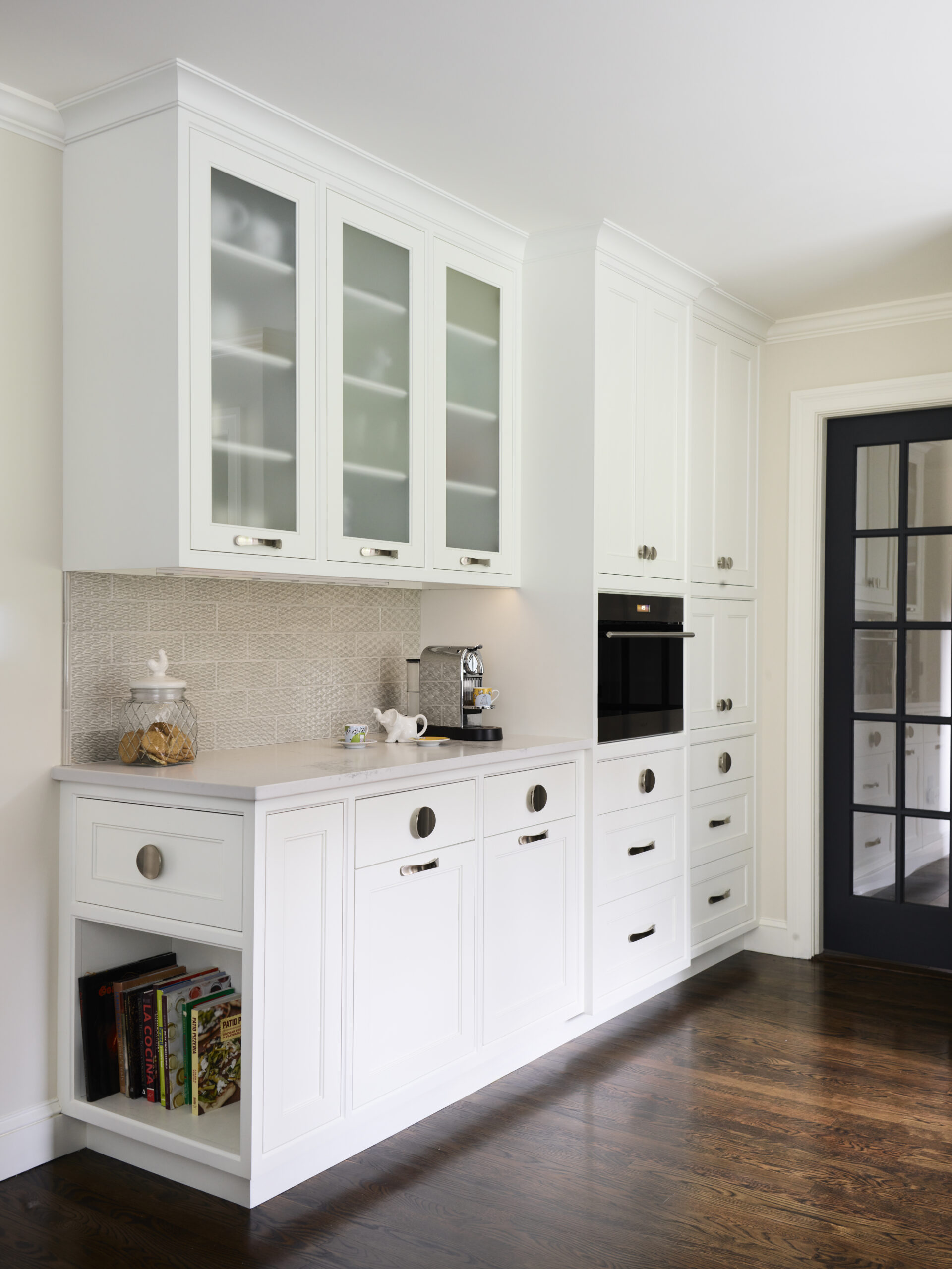 White kitchen cabinetry with glass upper doors, a built-in oven, countertop appliances, and a cookie jar on display reflect the refined touch of luxury interior design, curated by an experienced interior designer in Massachusetts.
