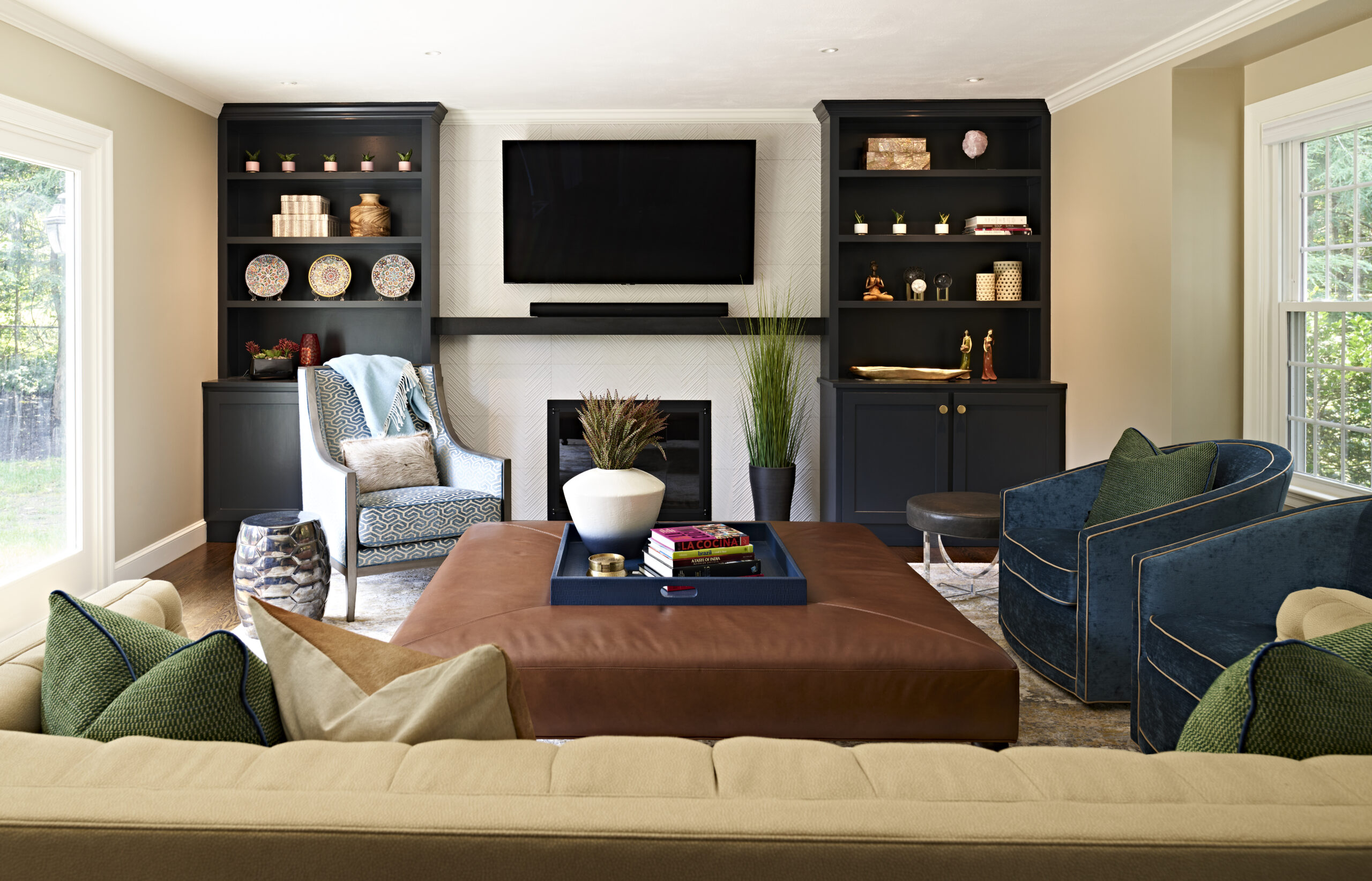 Modern living room with a beige sofa, blue armchairs, a large brown ottoman, built-in black shelves, a wall-mounted TV, and large windows letting in natural light—perfect for those who appreciate luxury interior design.