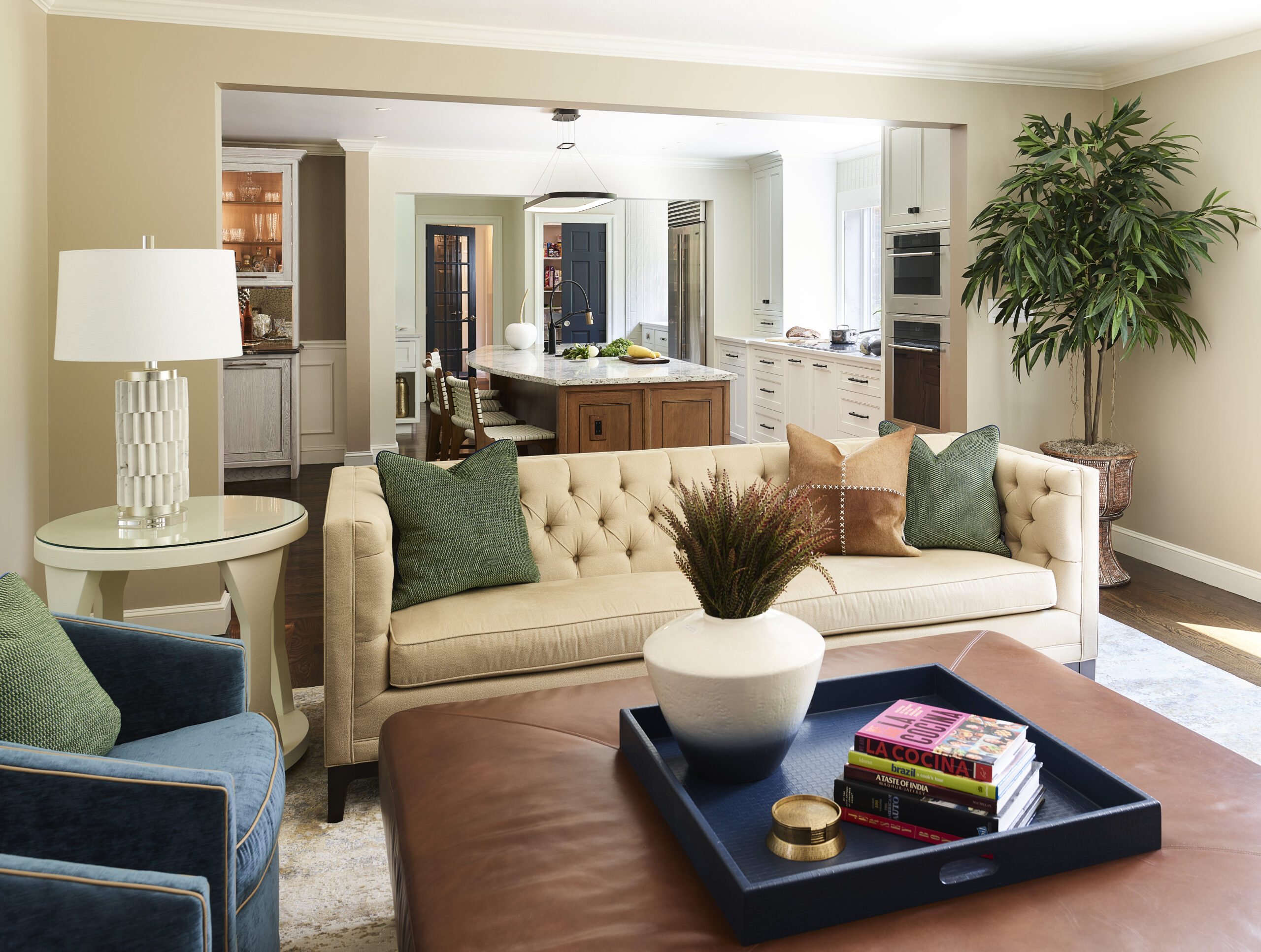 A living room with a beige tufted sofa, green and tan pillows, a blue armchair, and a brown ottoman with a tray showcases luxury interior design, opening into a kitchen area—a style often sought from interior designers in Massachusetts.