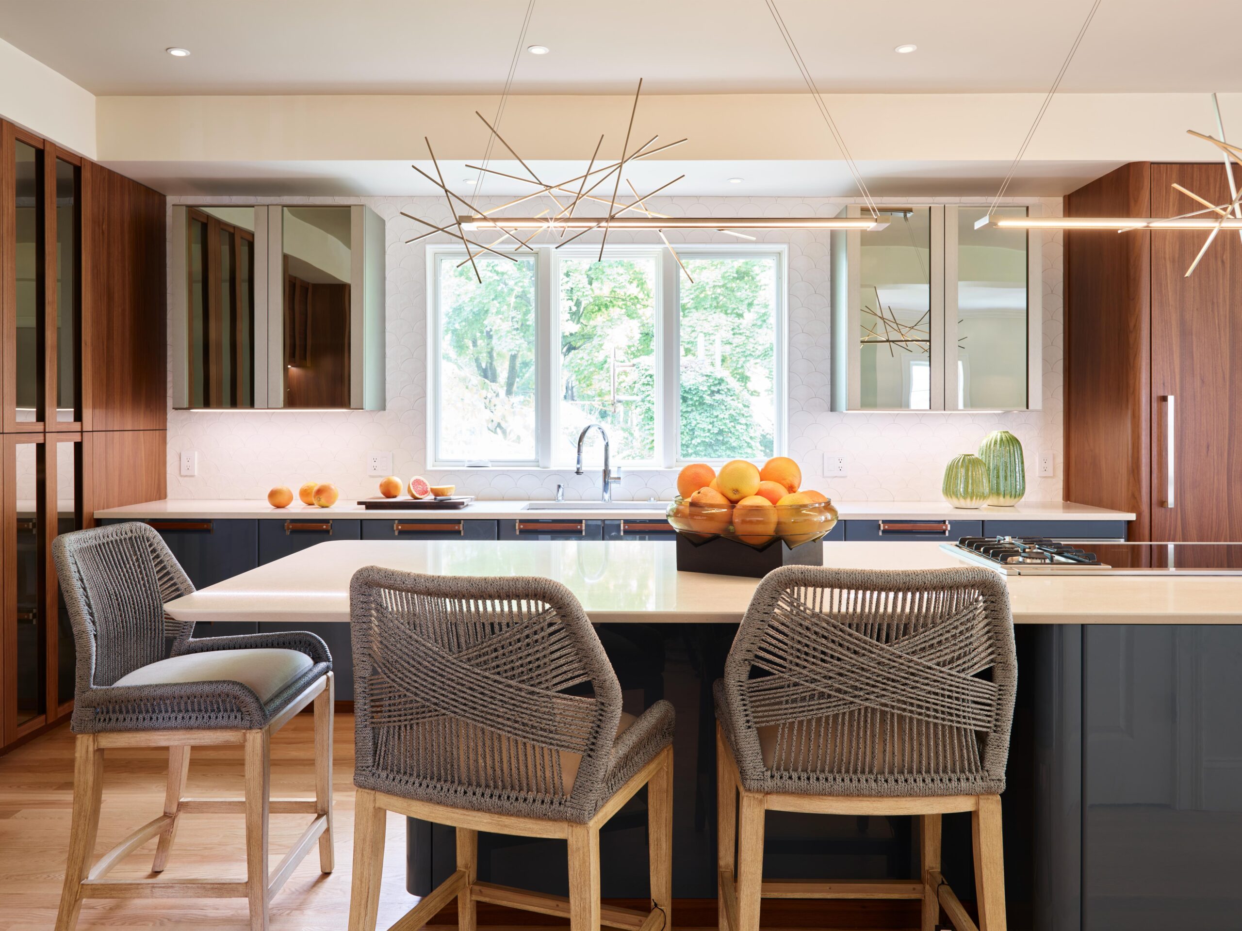 Modern kitchen interior design with wood cabinets, a large island featuring woven barstools, contemporary pendant lights, fruit bowls, and a window overlooking greenery.