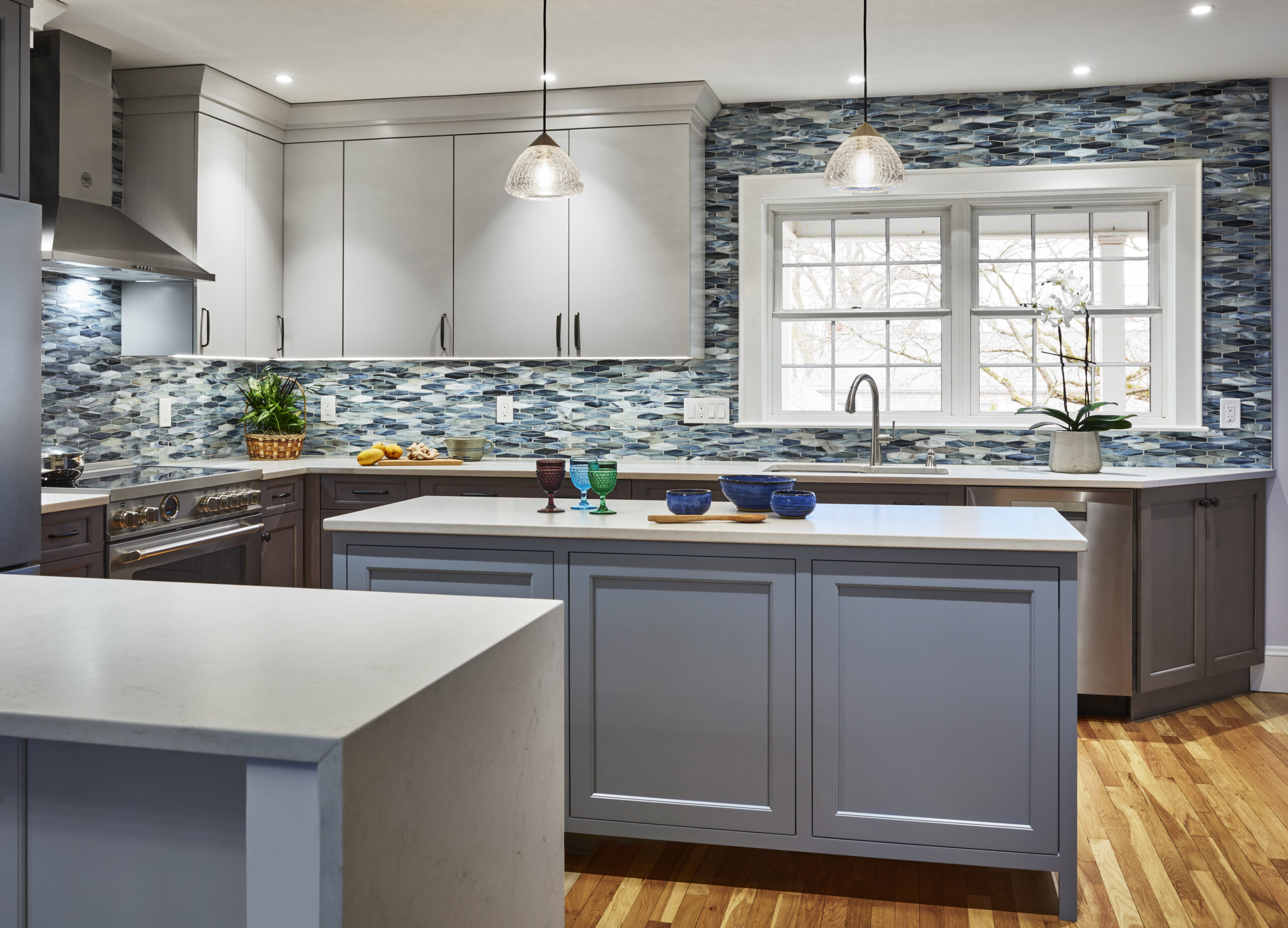 Modern kitchen with light gray cabinets, white countertops, blue mosaic tile backsplash, center island, pendant lights, and hardwood floors. Styled by an interior designer Massachusetts, it captures a touch of luxury interior design with elegant details and accents.
