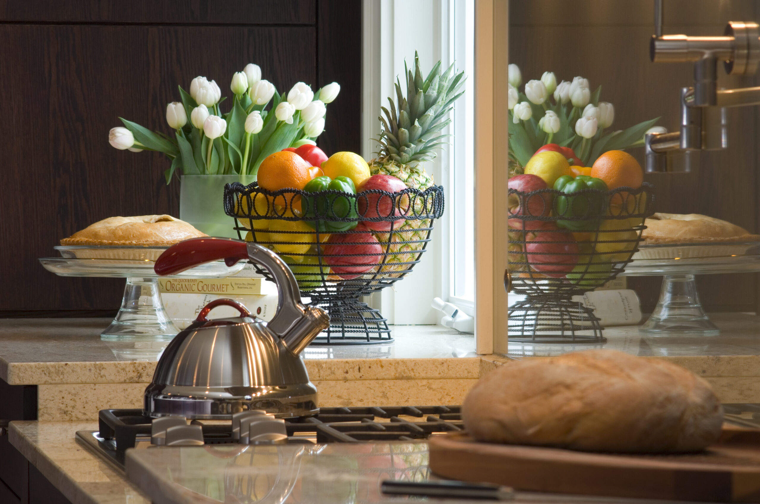 A kitchen countertop styled with luxury interior design features a kettle on the stove, a loaf of bread on a cutting board, a fruit basket, two pies, and white tulips in a vase near the window.