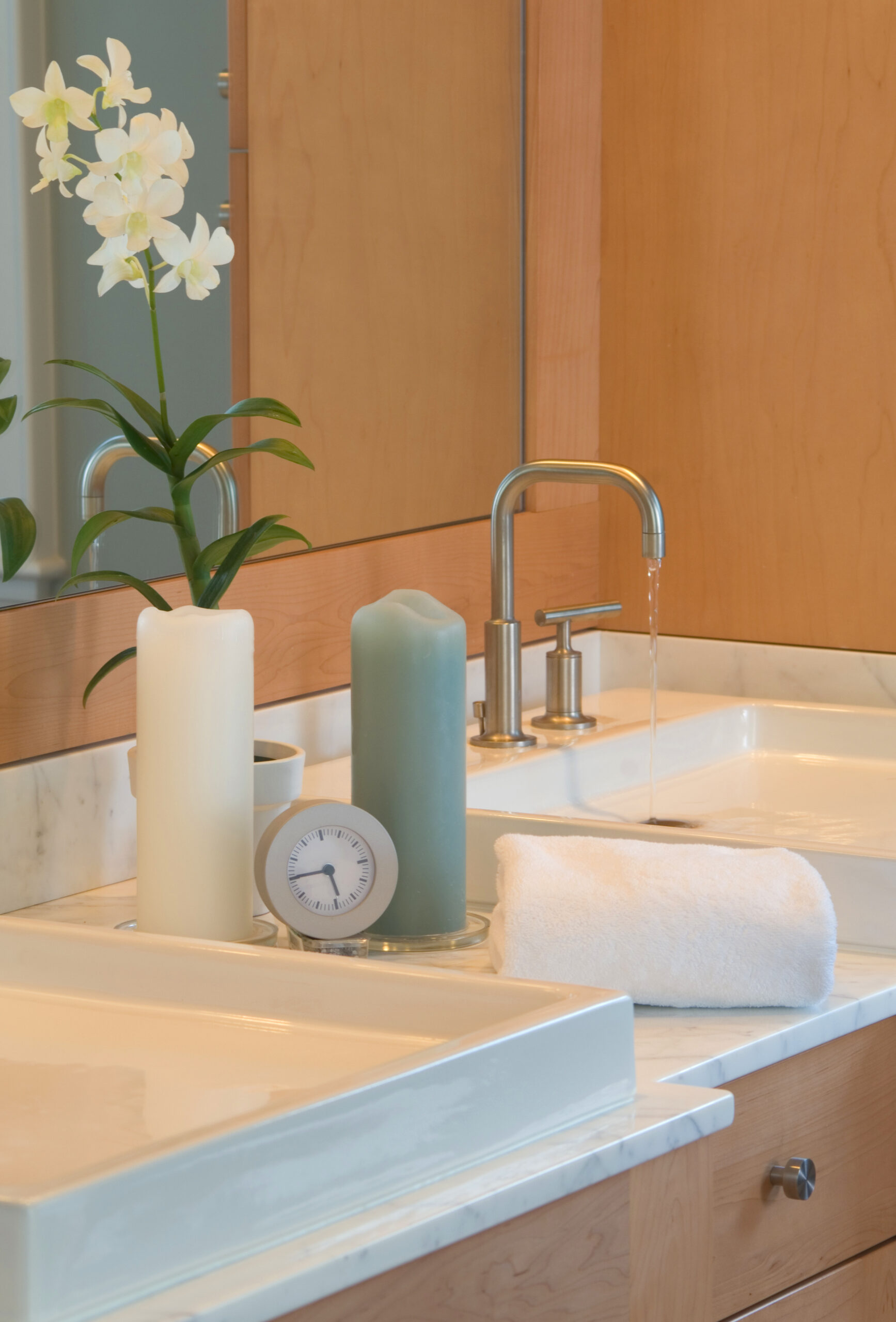 Modern bathroom sink with running water, a white rolled towel, two pillar candles, a small clock, an orchid plant, and wooden cabinets—perfect for those seeking luxury interior design inspired by top interior designers in Massachusetts.