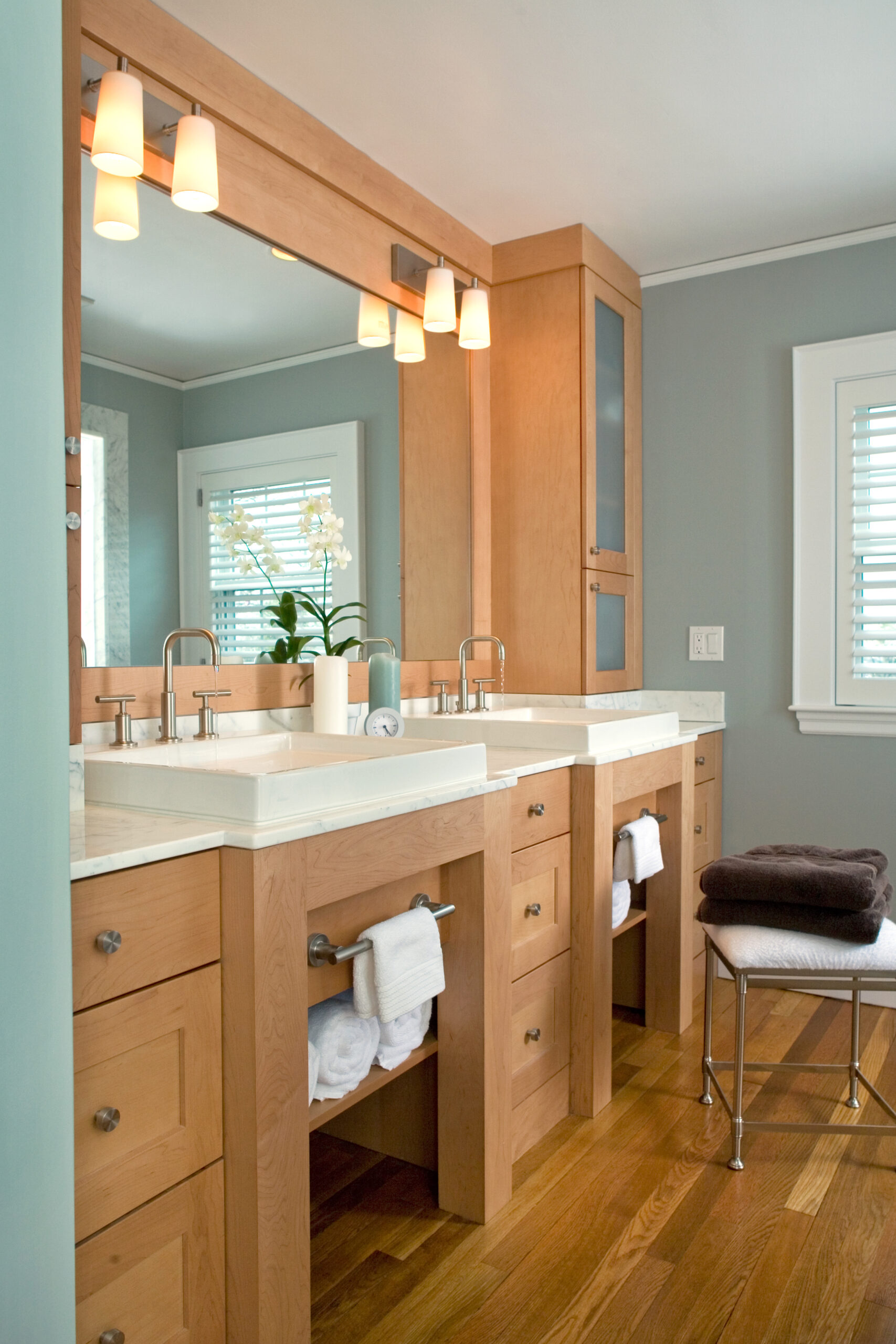 Modern bathroom with two wooden vanities, double sinks, large mirror, wall-mounted lights, folded towels, a small orchid plant, and a window with white blinds—a true showcase of luxury interior design.