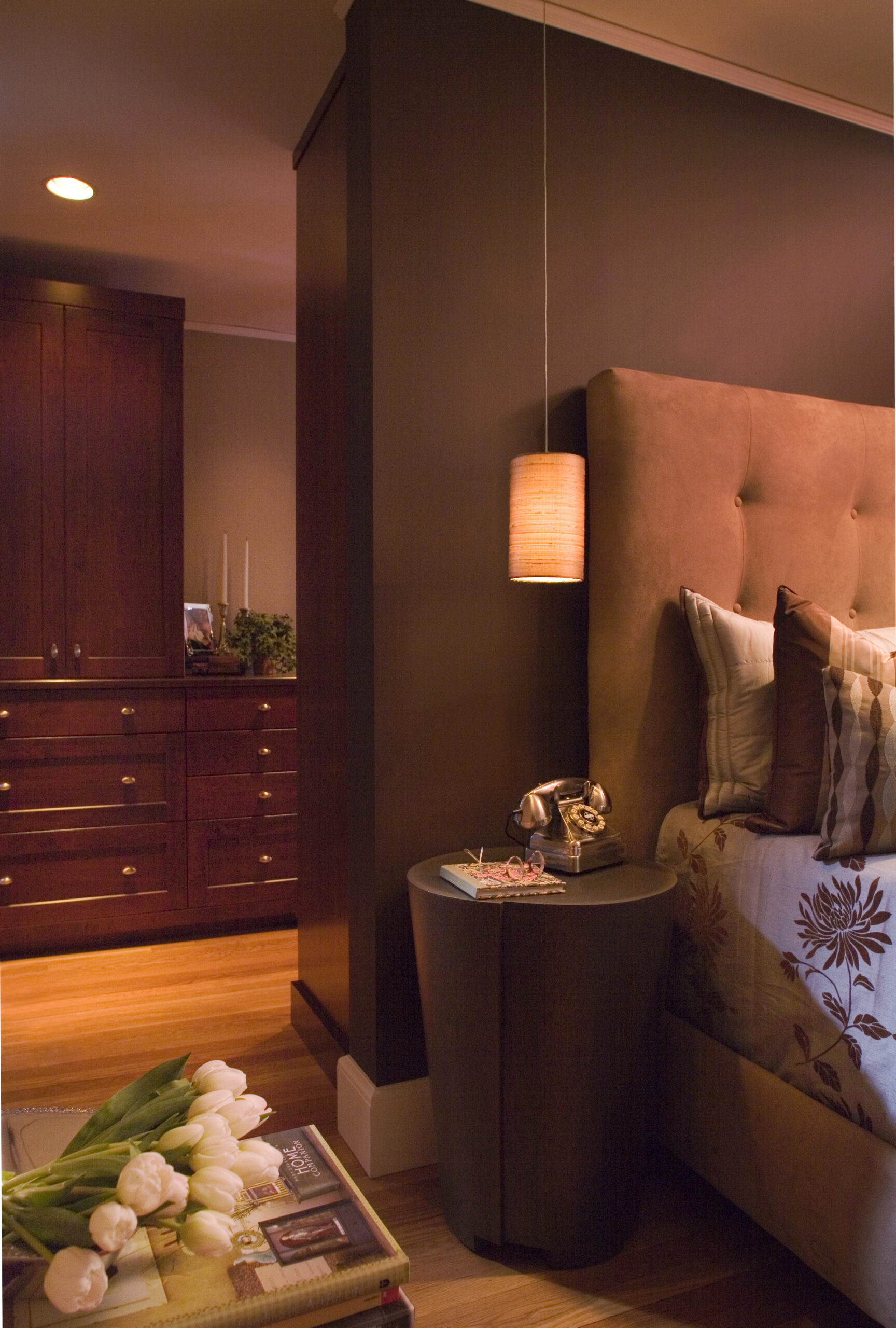 A bedside table with a retro phone, glasses, and pendant light beside a bed with patterned pillows showcases luxury interior design; in the background, a dresser and tulips add elegance to this thoughtfully styled Massachusetts space.
