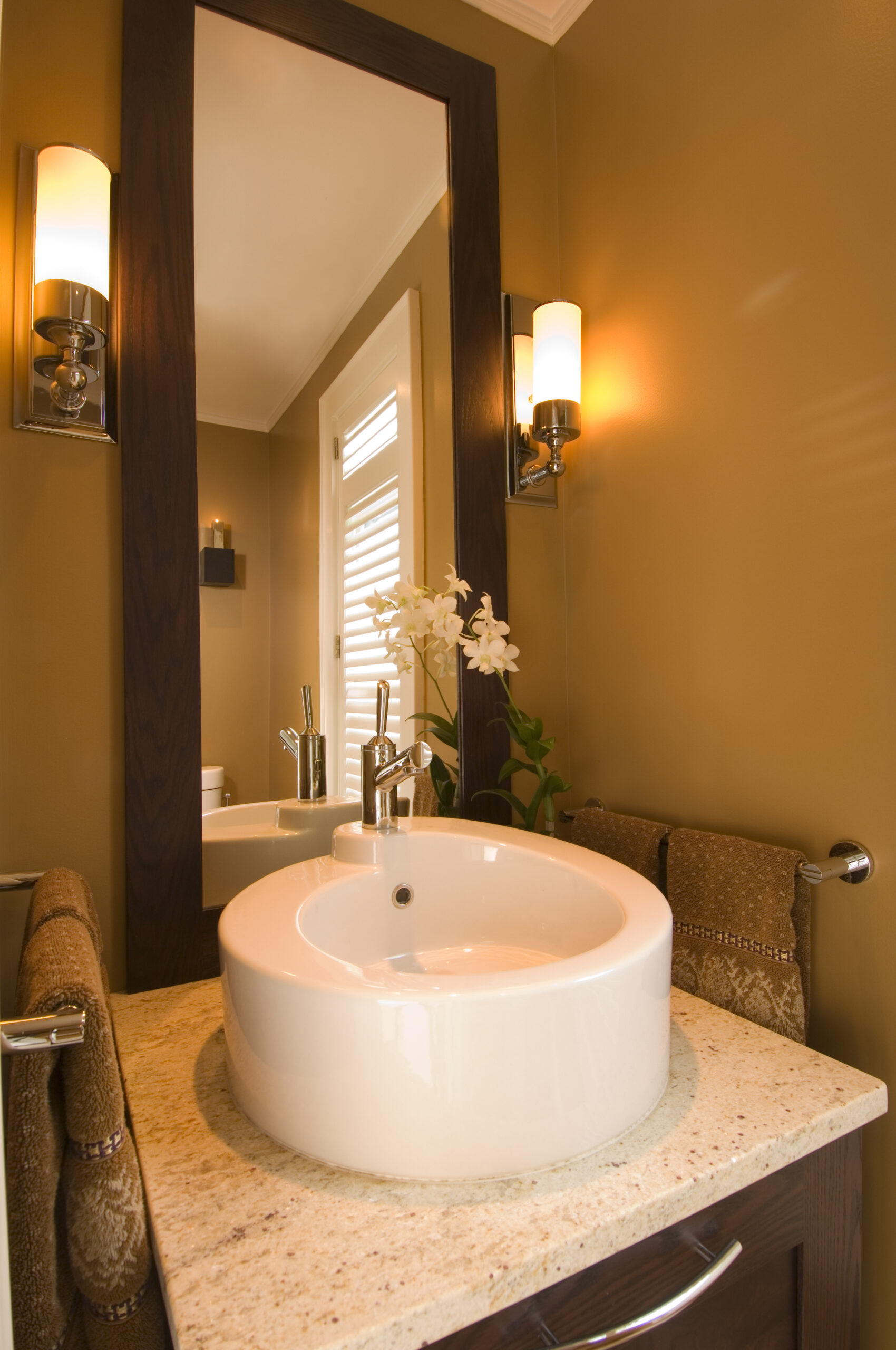 Modern bathroom vanity with a round white sink, granite countertop, large mirror, wall sconces, towels, and a white orchid in a vase—perfect for those seeking luxury interior design details.