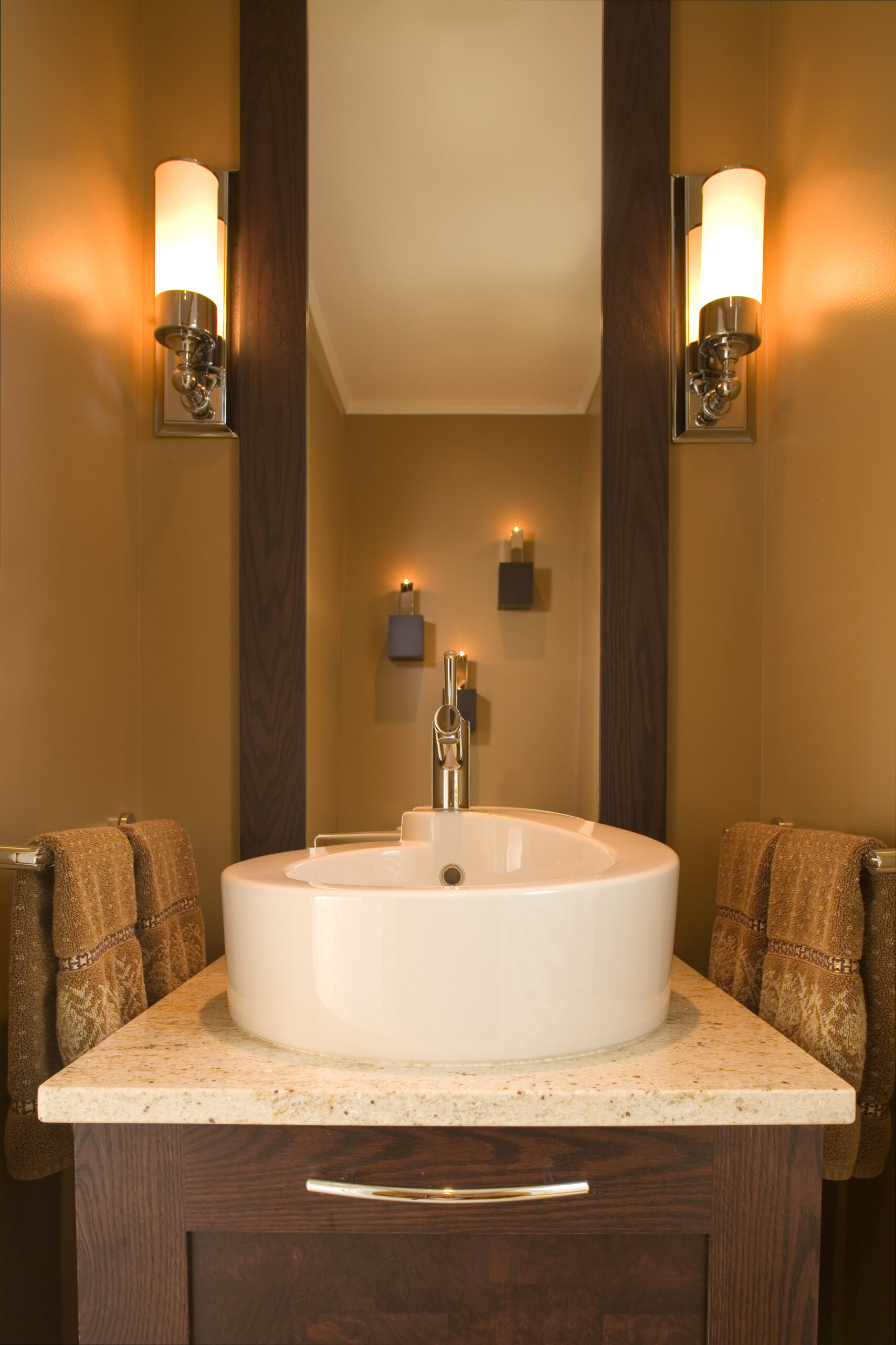 Modern bathroom with a round white sink, chrome faucet, brown wood vanity, and tan walls—showcasing Luxury Interior Design touches by an expert Interior Designer Massachusetts. Wall sconces, brown hand towels, and candles complete the serene space.