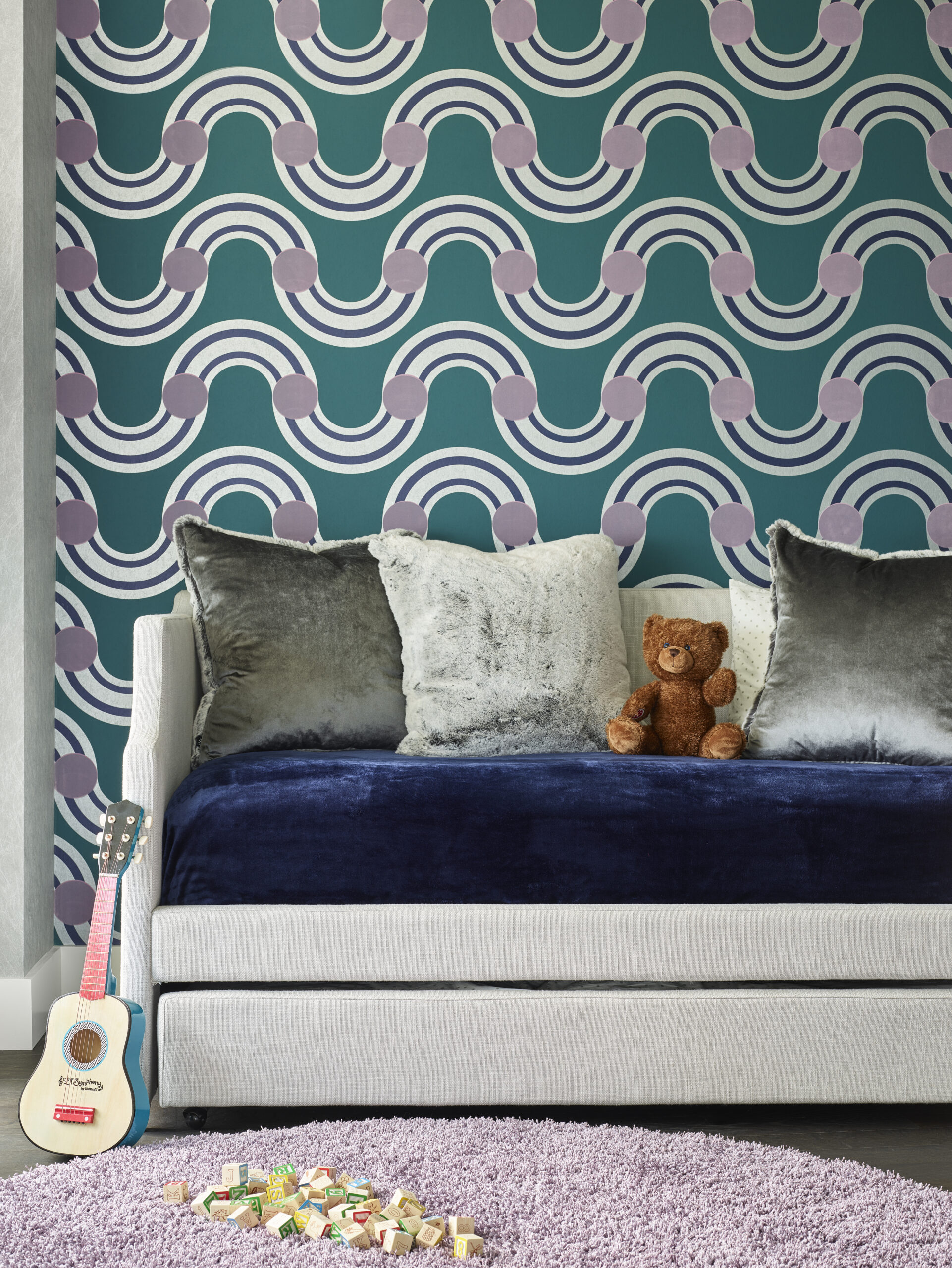 A small couch with velvet pillows and a teddy bear sits against a bold, geometric patterned wall. Interior Designer Massachusetts touches show in the details, from the toy guitar to wooden blocks on the purple rug in front.