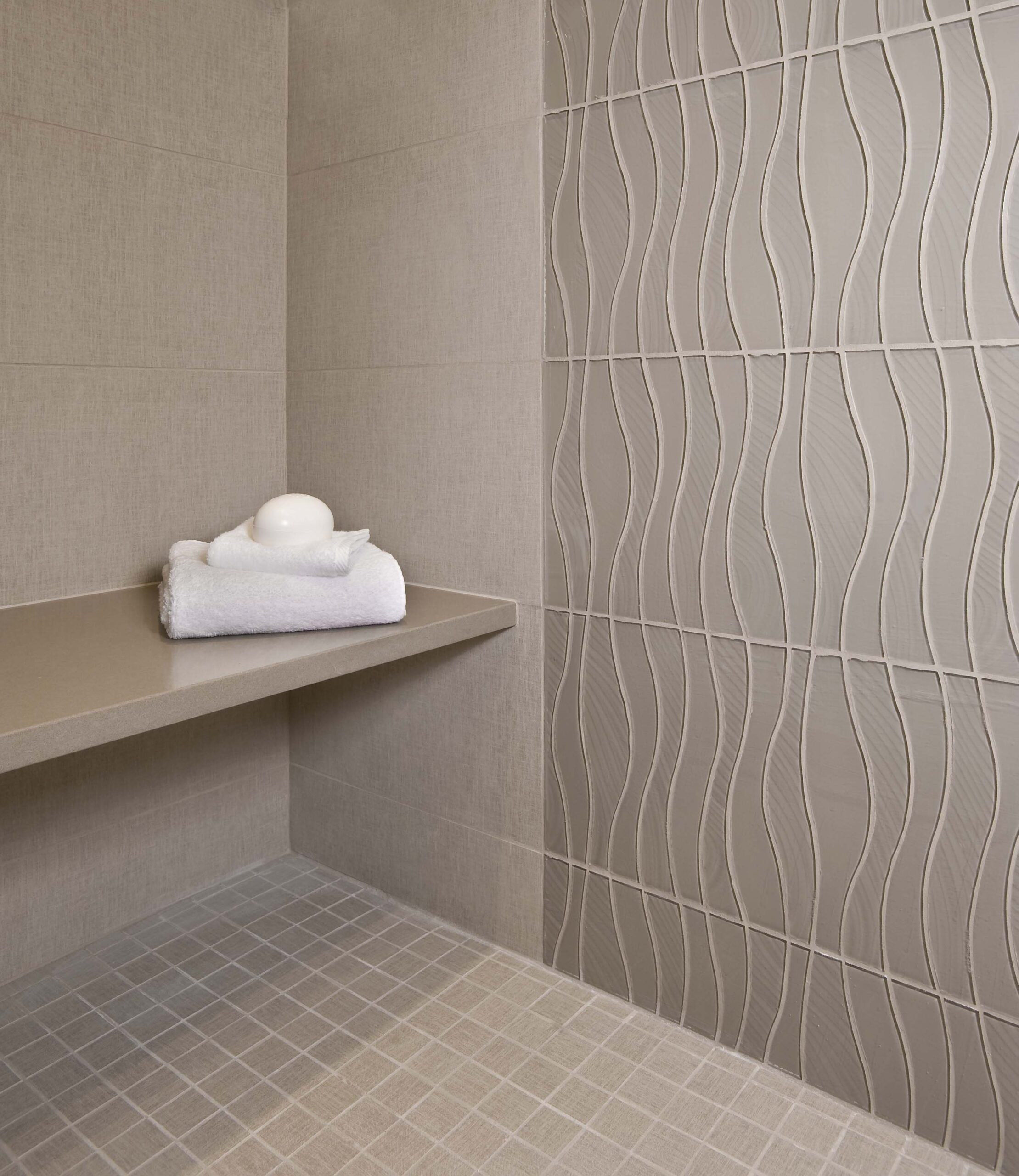 A modern shower corner with beige tiles, a built-in bench, a folded white towel, and a bar of soap; wall tiles feature a wavy textured pattern, showcasing luxury interior design by an expert interior designer in Massachusetts.