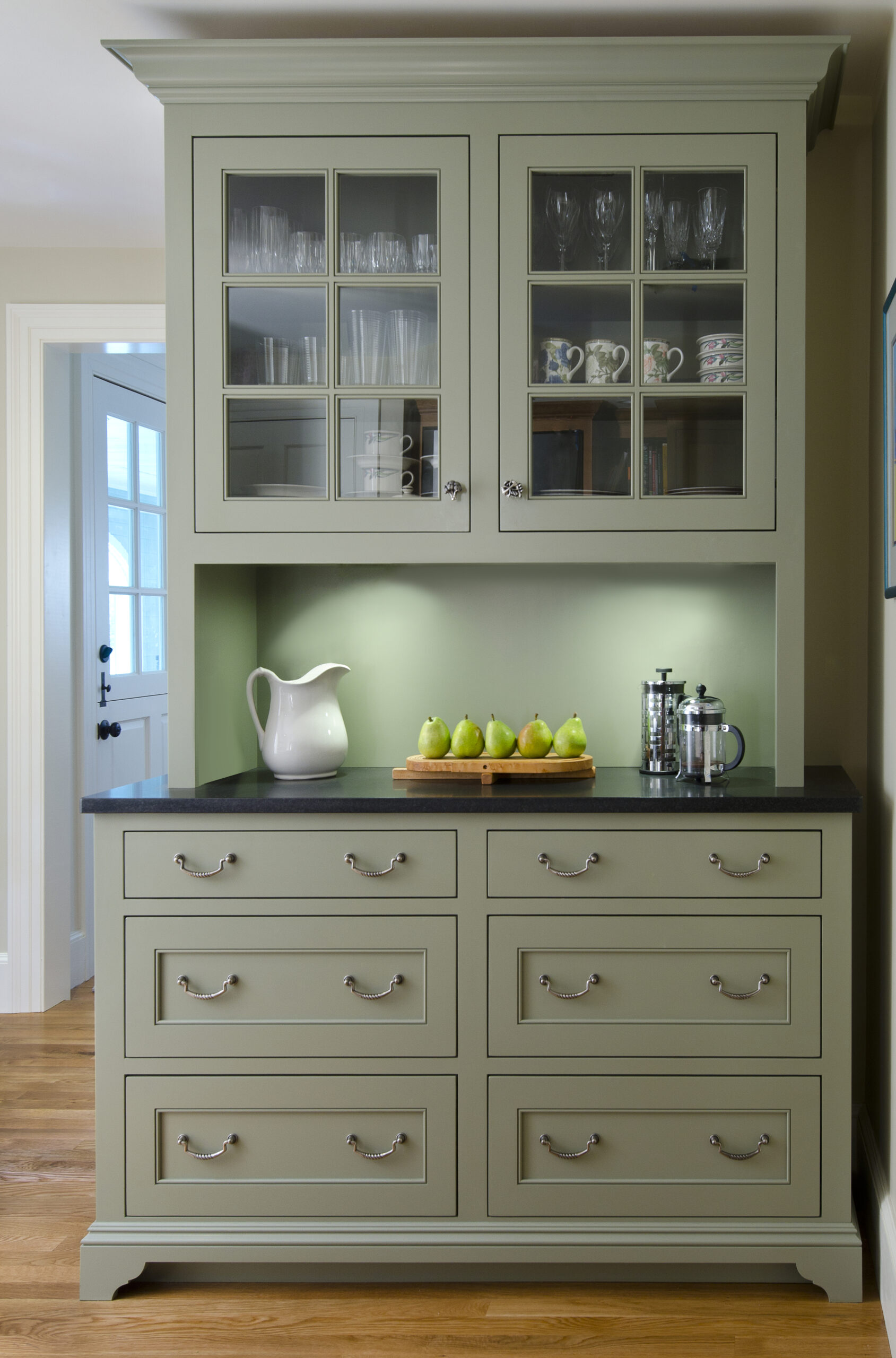 A sage green kitchen hutch with glass-front upper cabinets, drawers below, and a black countertop displaying a white pitcher, a row of pears, and a French press—perfect for those inspired by luxury interior design.