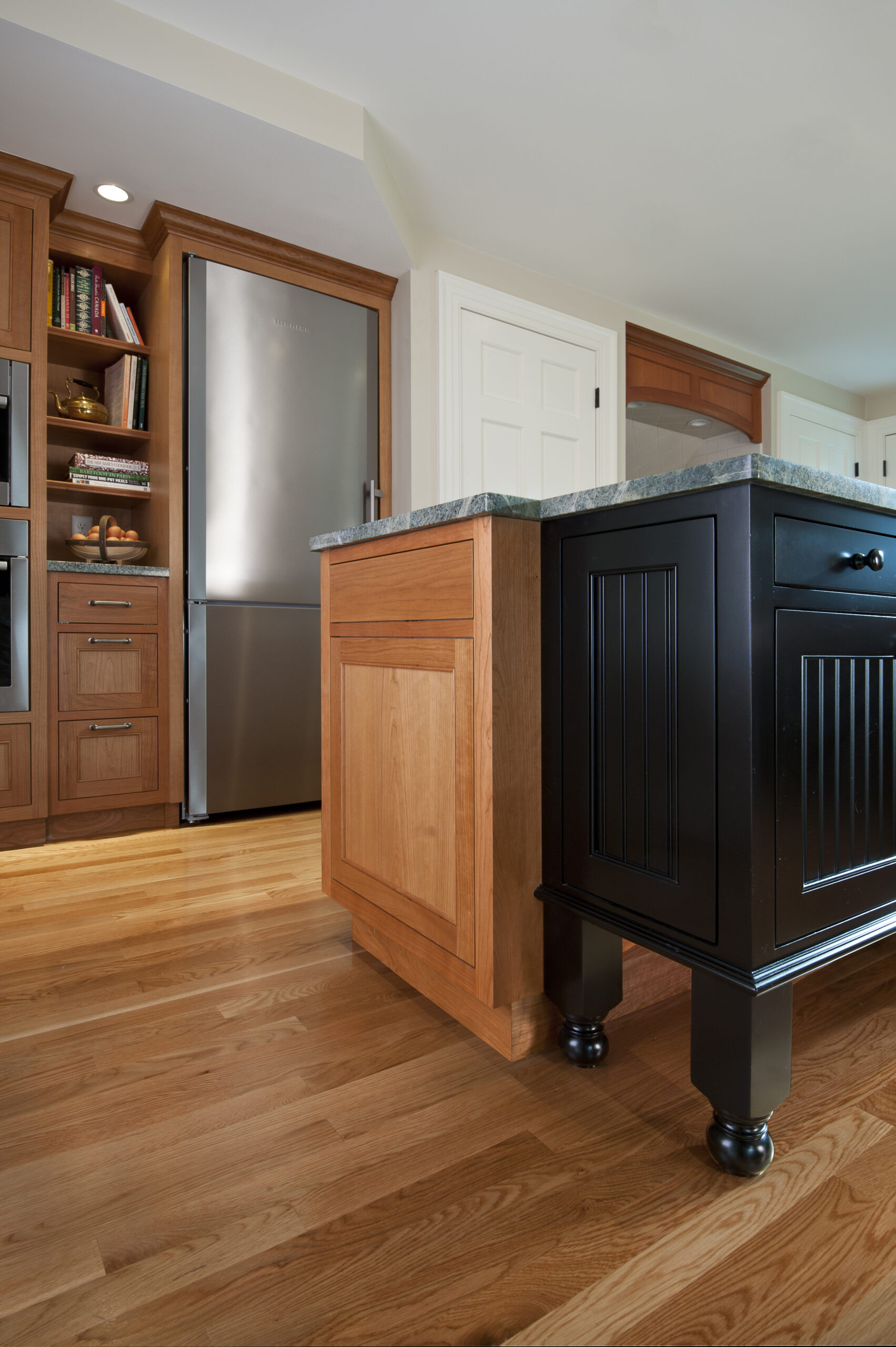 Modern kitchen with wood cabinets, stainless steel refrigerator, a black island with panel detailing, and hardwood flooring—perfectly crafted by a leading interior designer Massachusetts for luxury interior design enthusiasts.