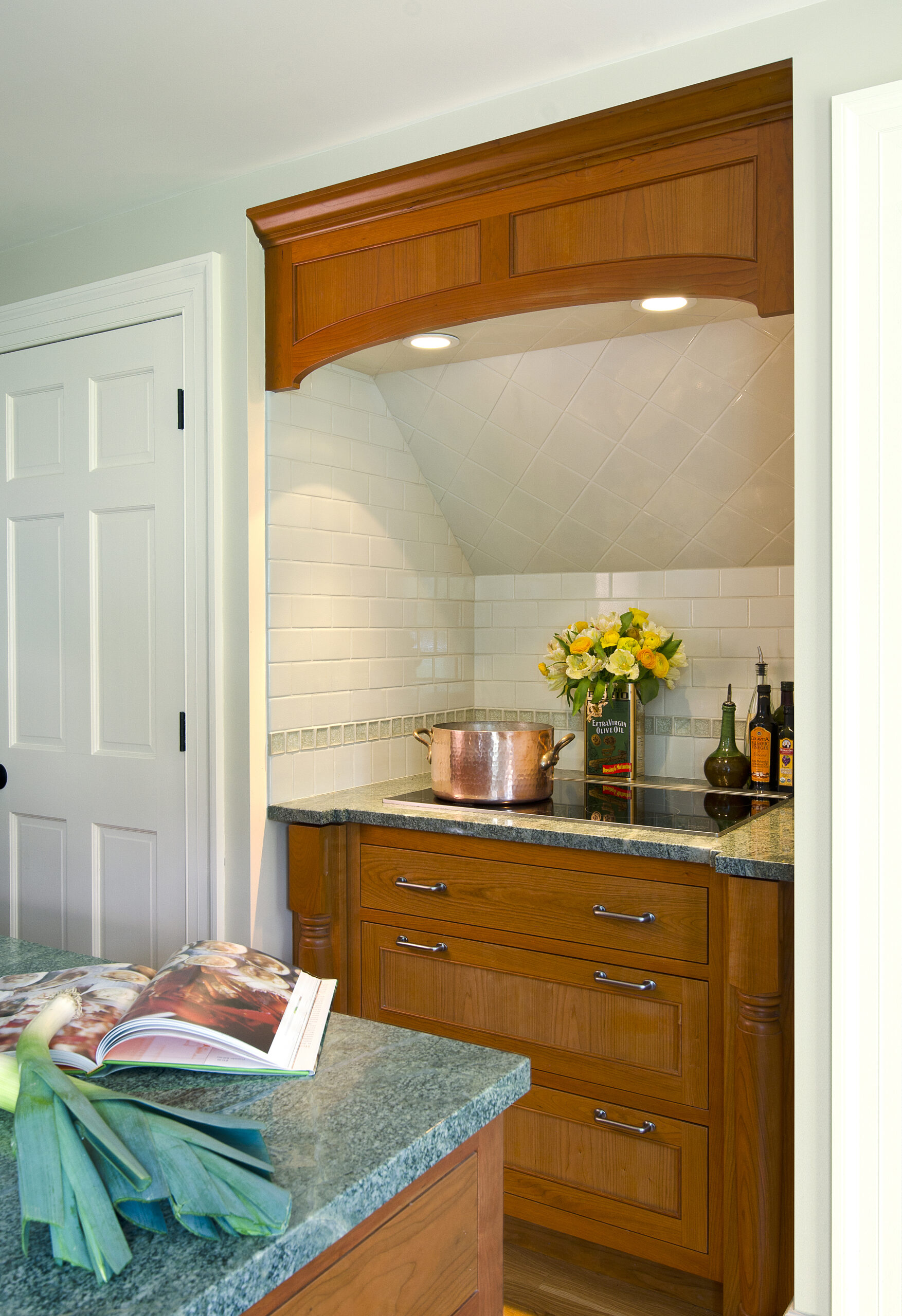 A kitchen nook with wooden cabinets, granite countertops, a copper pot on the stove, a vase of yellow flowers, and bottles on the tiled backsplash shelf exudes Luxury Interior Design perfect for any discerning homeowner.
