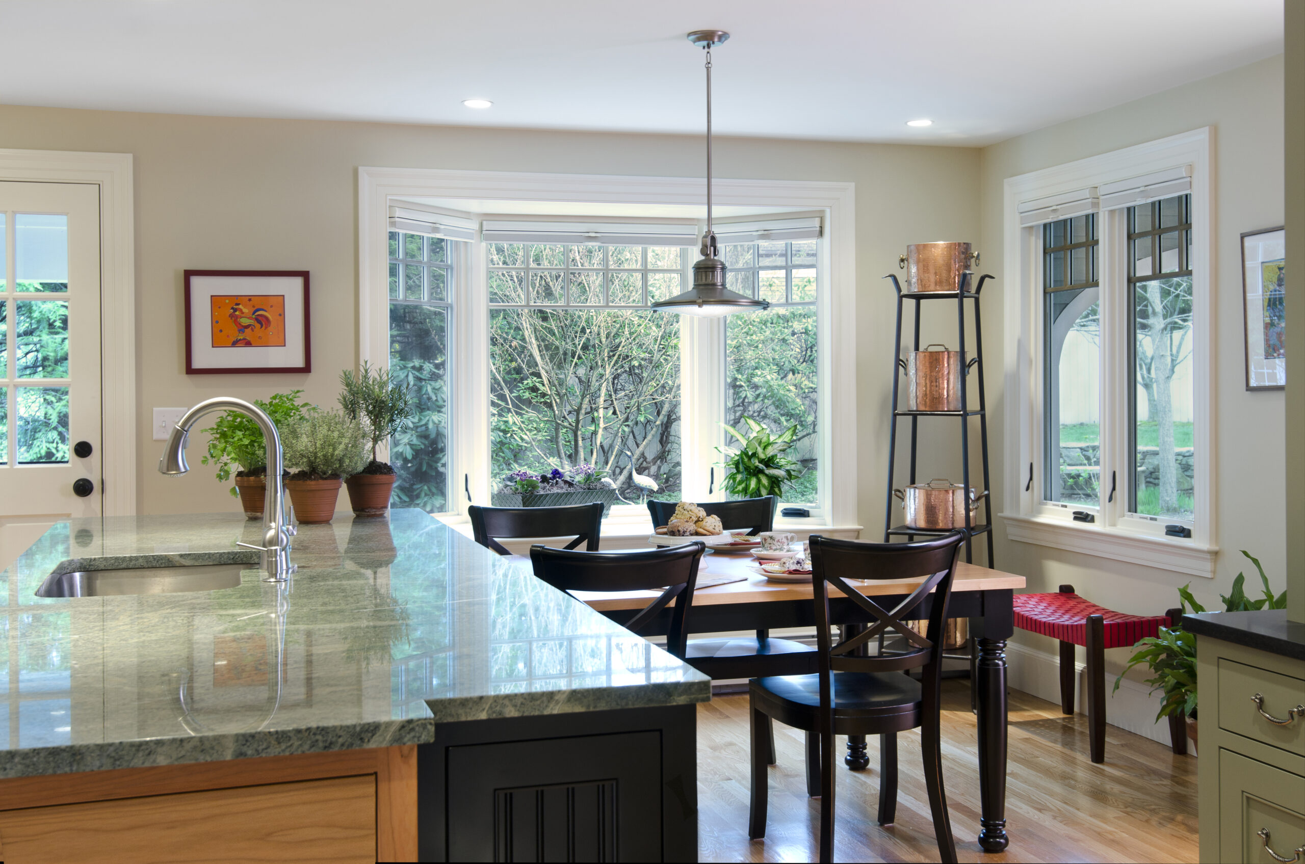 Bright kitchen and dining area with a marble island, potted plants, wooden table with chairs, large windows, and a view of greenery outside—perfectly styled for luxury interior design by an expert interior designer in Massachusetts.