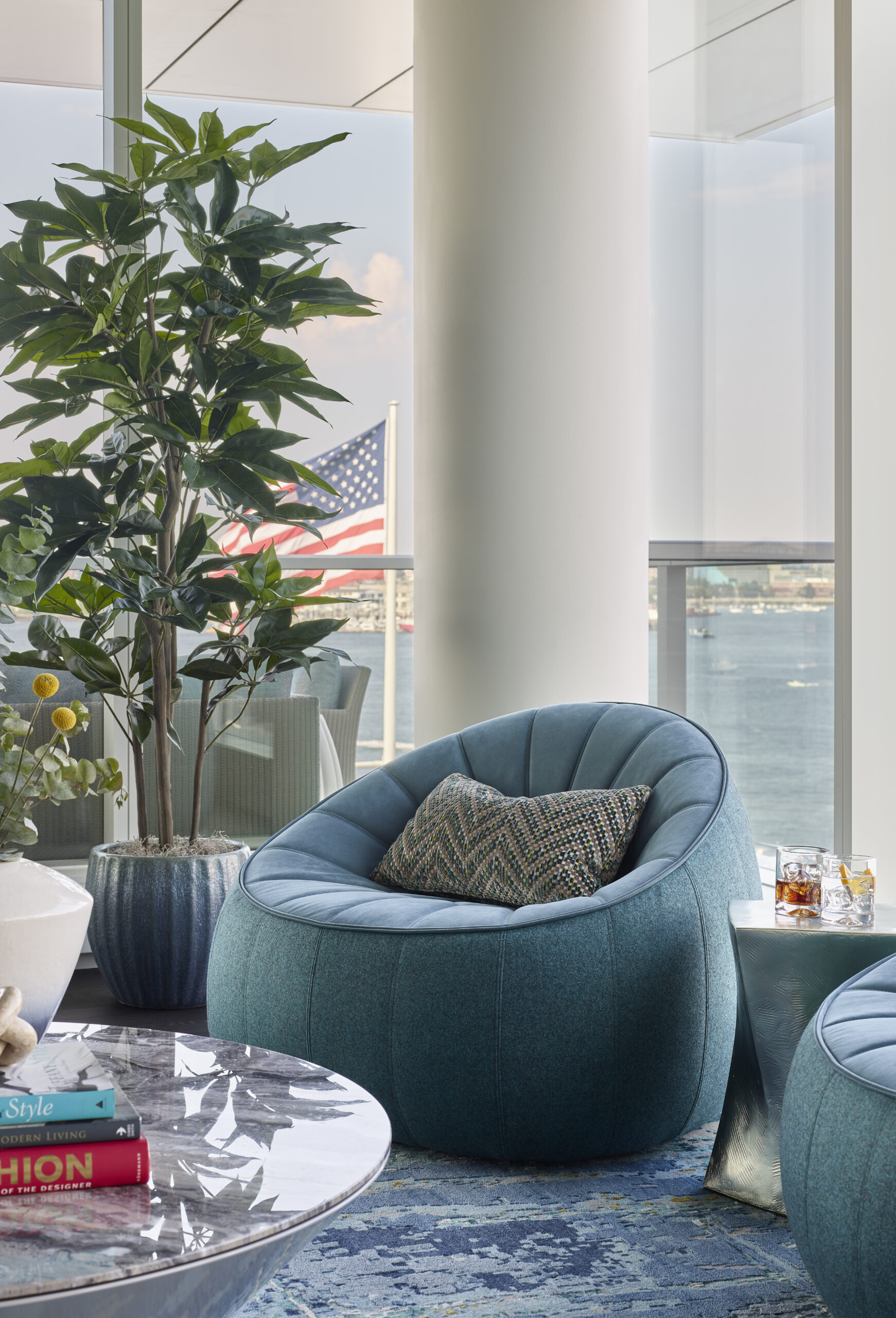A modern lounge area with a teal cushioned chair, patterned pillow, marble coffee table, potted plants, and a large window displaying water and an American flag—an inviting space inspired by Luxury Interior Design.