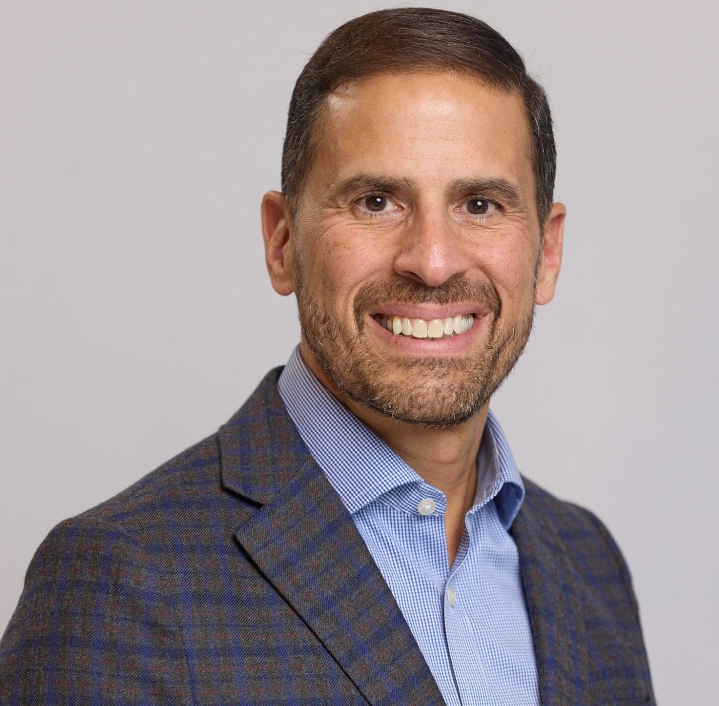 Smiling man with short brown hair and beard, dressed in a blue shirt and checked blazer, posed against a plain light background—perfectly embodying the polished style of a Luxury Interior Design expert in Massachusetts.