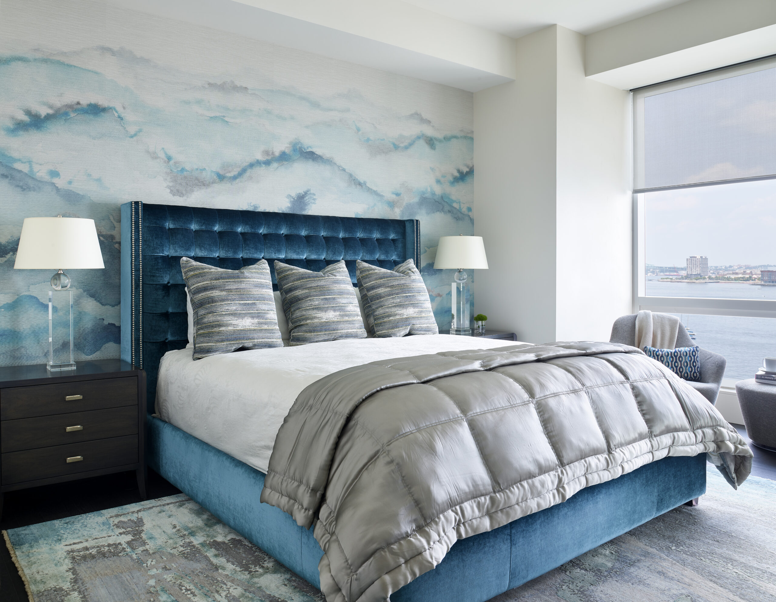 Modern bedroom with a blue upholstered bed, gray bedding, patterned accent pillows, two bedside tables with lamps, and a large window with a city view—perfect for lovers of luxury interior design.