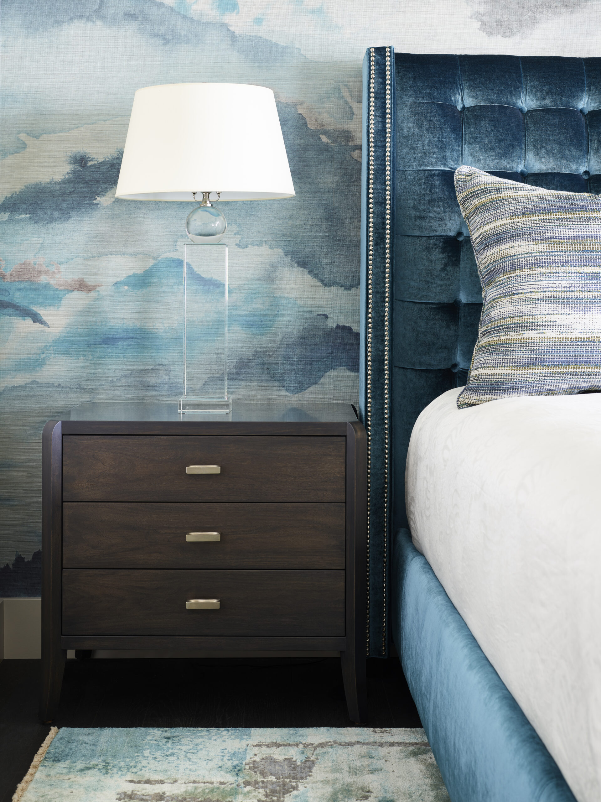 A modern bedside table with three drawers and a glass lamp sits next to a blue upholstered bed with a striped pillow, showcasing Luxury Interior Design against a blue watercolor-patterned wall.