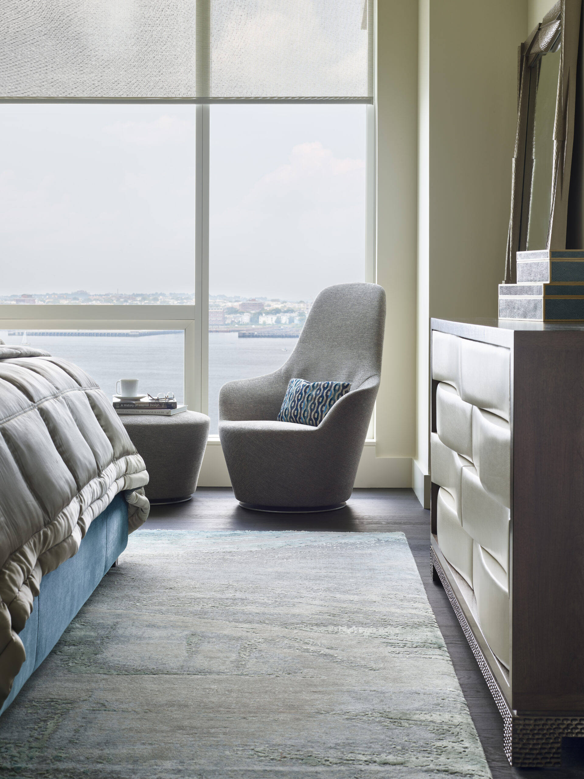 A modern bedroom designed with luxury interior design features a gray armchair, ottoman, and accent pillow by a large window overlooking water, next to a bed and dresser.