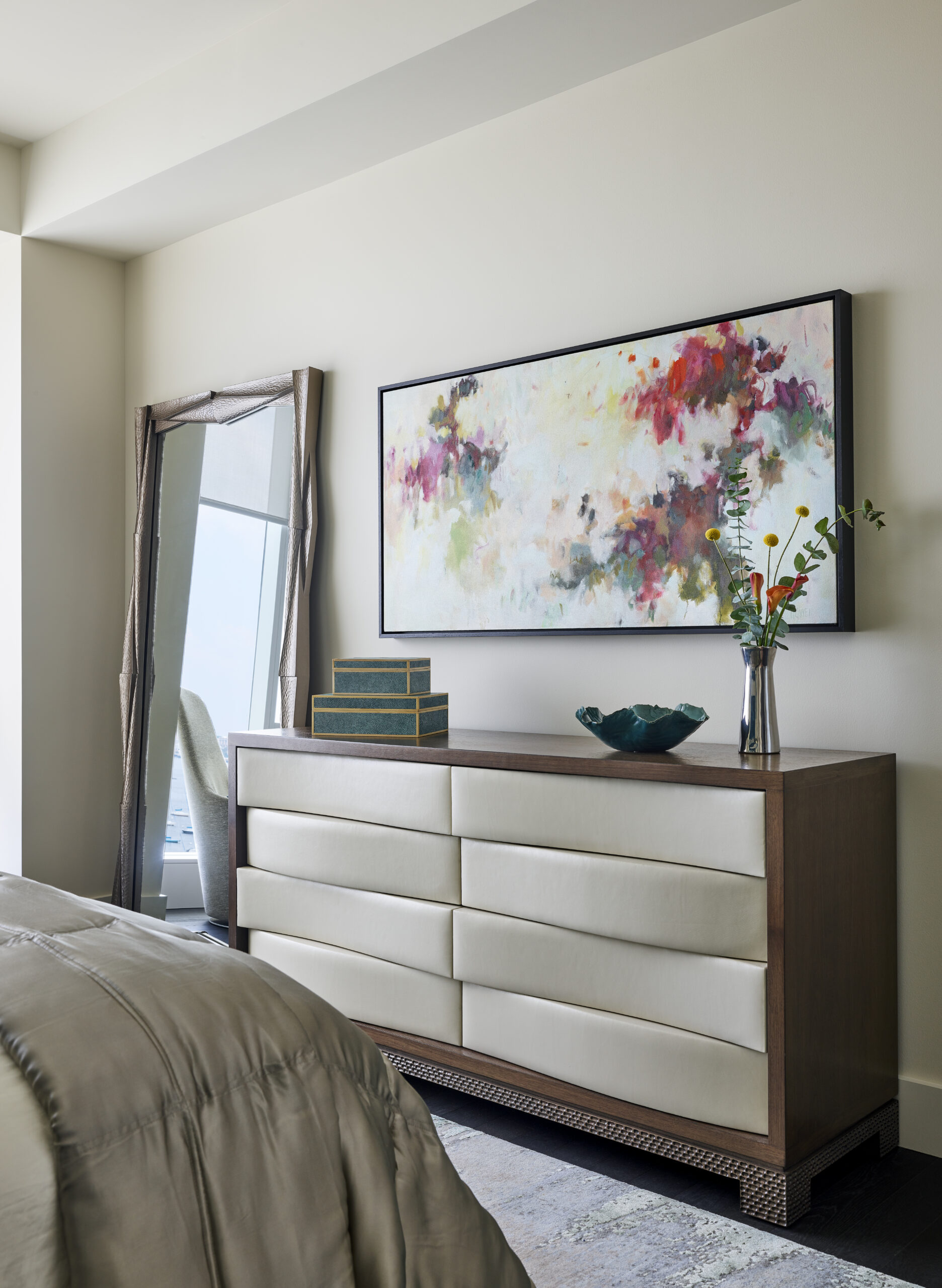 A modern bedroom dresser styled with a decorative bowl, stacked boxes, vase of flowers, a large abstract painting above, and a tall mirror—perfect inspiration for Luxury Interior Design by an expert Interior Designer in Massachusetts.