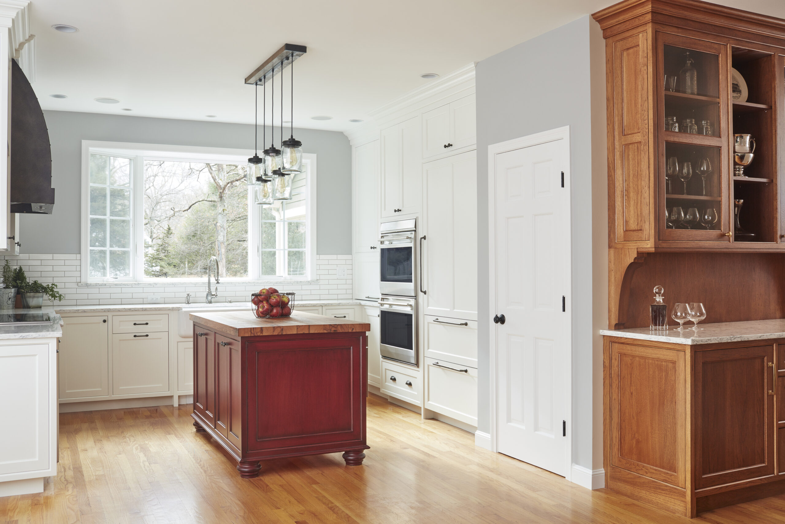 Modern kitchen with white cabinets, a red island, stainless steel appliances, wood floors, and a window overlooking trees. Designed by an Interior Designer Massachusetts, it features a glass-front cabinet on the right displaying elegant glassware.