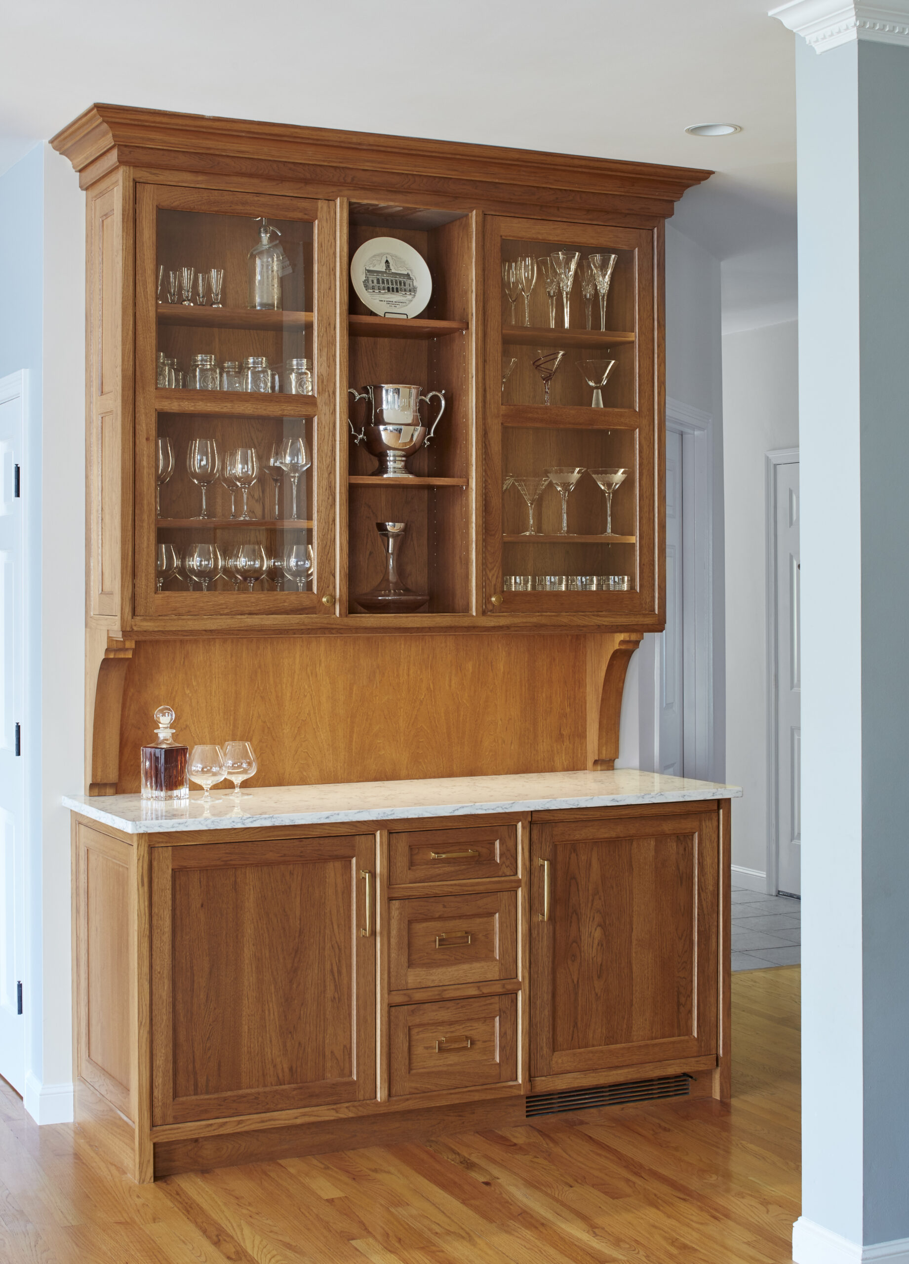A wooden cabinet with glass doors displays glassware and trophies above a marble countertop with a decanter and glasses, set against a light blue wall—a perfect example of luxury interior design.