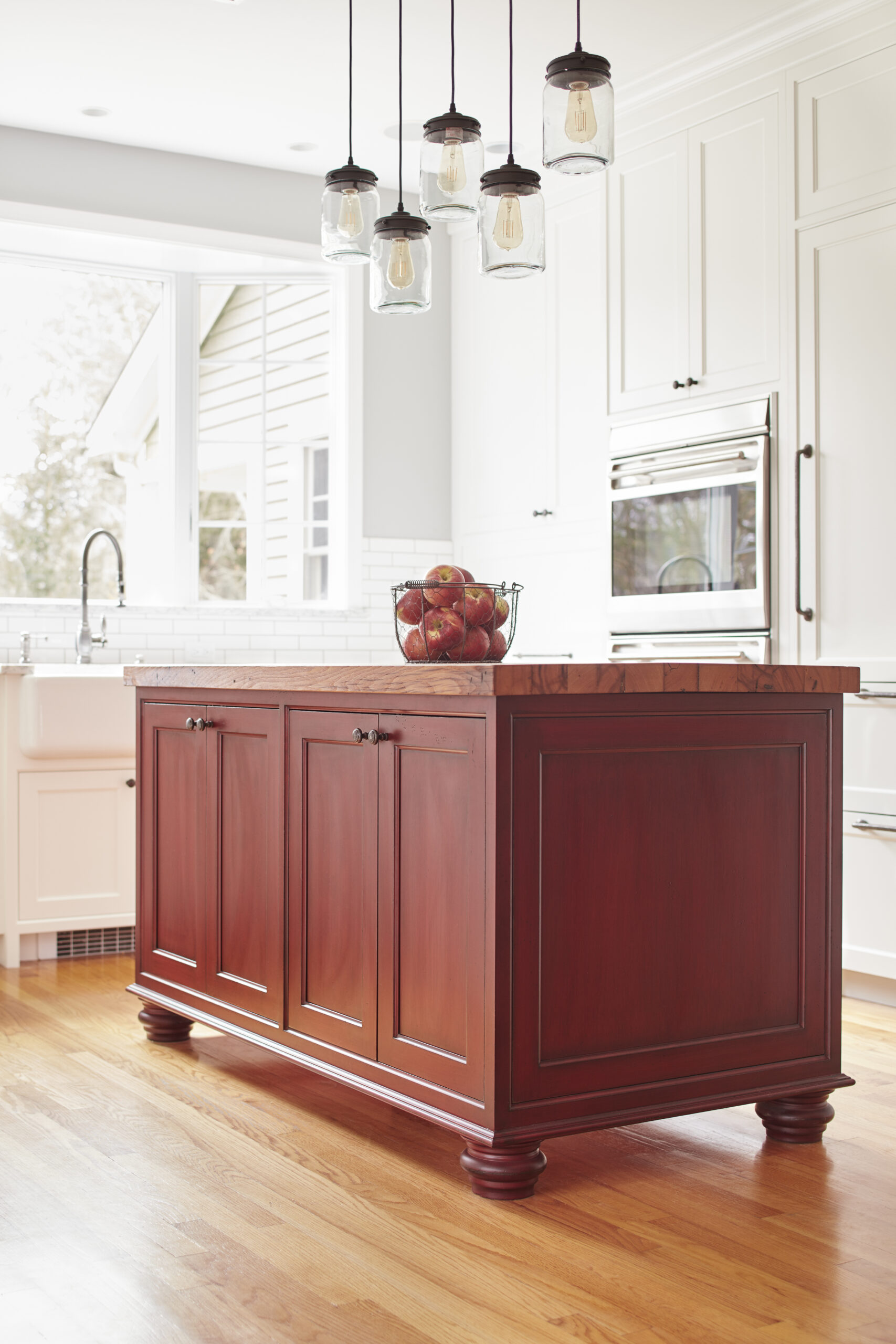 A kitchen with a wooden island topped with a bowl of apples, pendant lights above, white cabinets, an oven, and a sink near a large window showcases the elegance of luxury interior design.