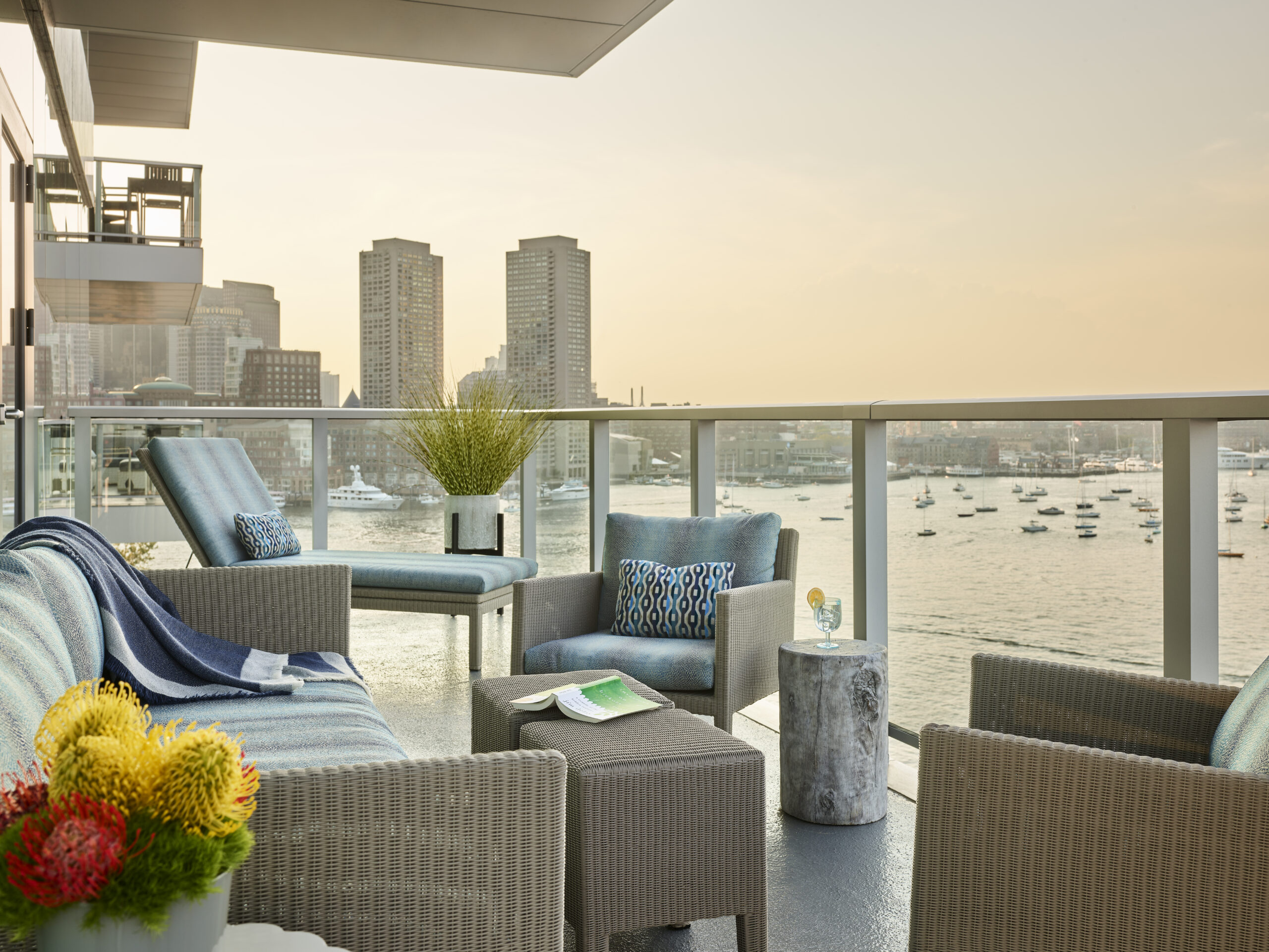 Contemporary outdoor patio with cushioned seating by an Interior Designer Massachusetts overlooks a harbor with moored boats and city buildings in the background at sunset.