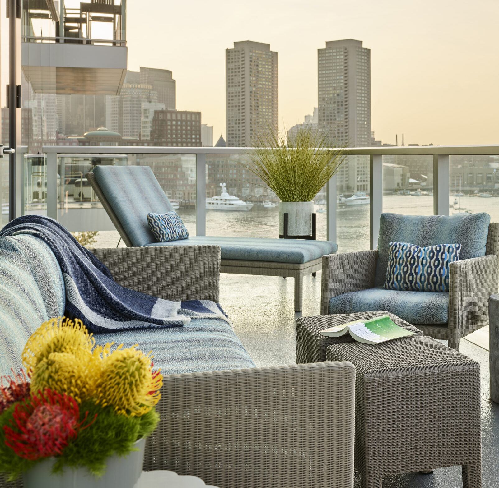 A modern balcony by an Interior Designer Massachusetts features wicker patio furniture, colorful cushions, a flower arrangement, and a city skyline with water and tall buildings—showcasing luxury interior design in every detail.