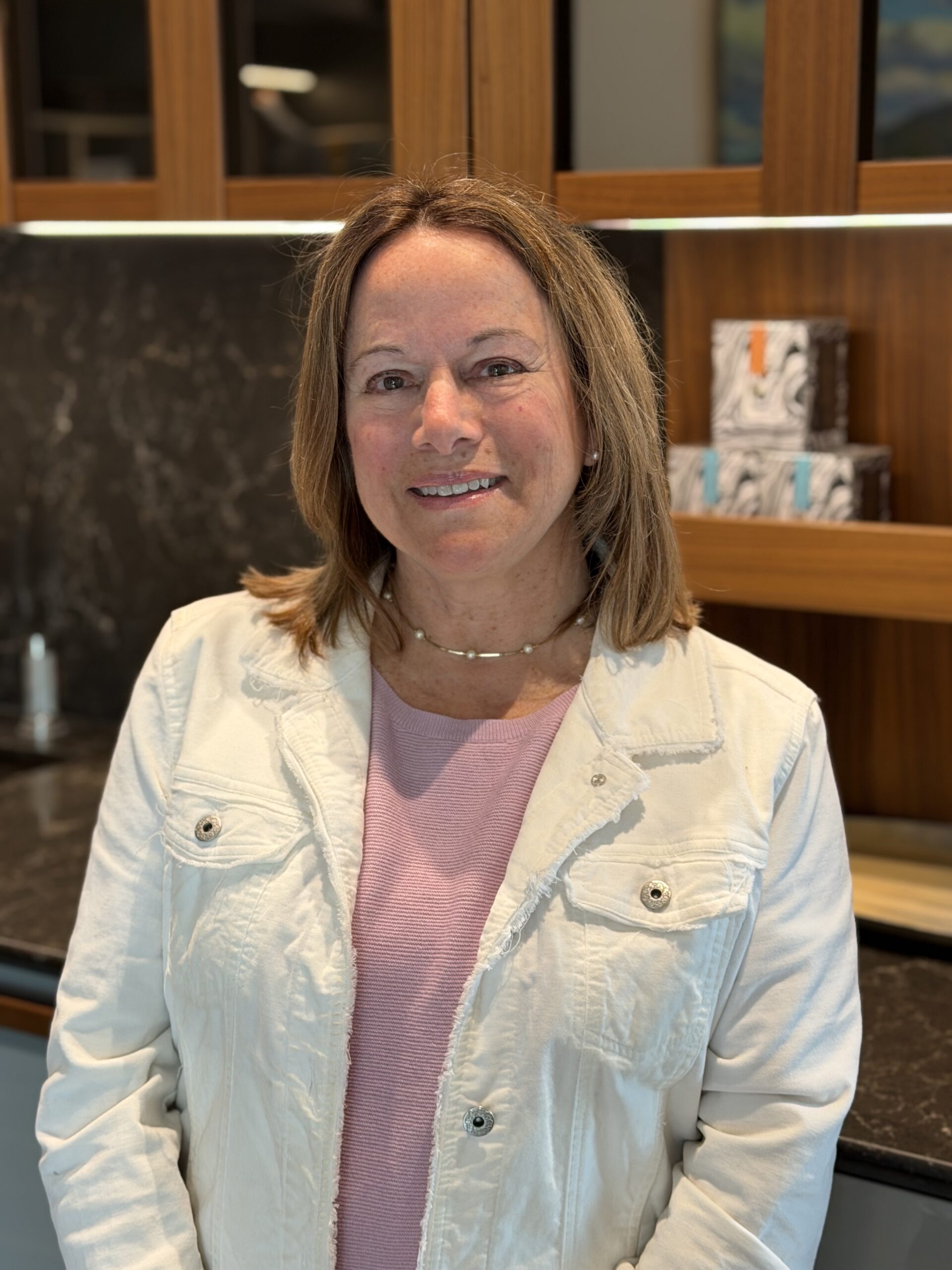 A woman with shoulder-length light brown hair wearing a white jacket and pink top stands indoors in a kitchen with wooden cabinets and a dark countertop, showcasing luxury interior design by an Interior Designer Massachusetts.