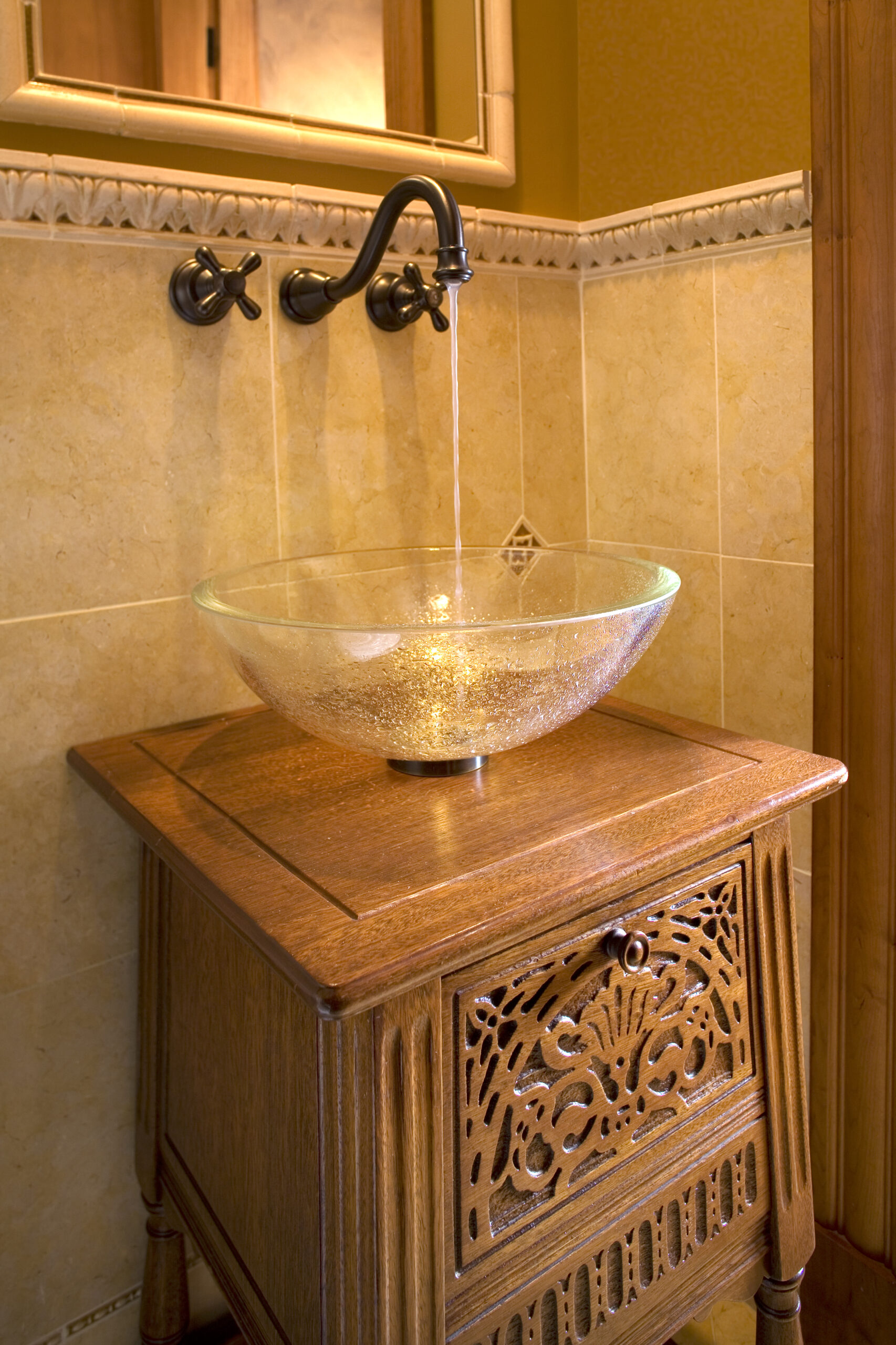 A glass vessel sink with water running from a wall-mounted faucet sits on an ornate wooden vanity in a bathroom with beige tiled walls, exemplifying the essence of luxury interior design.
