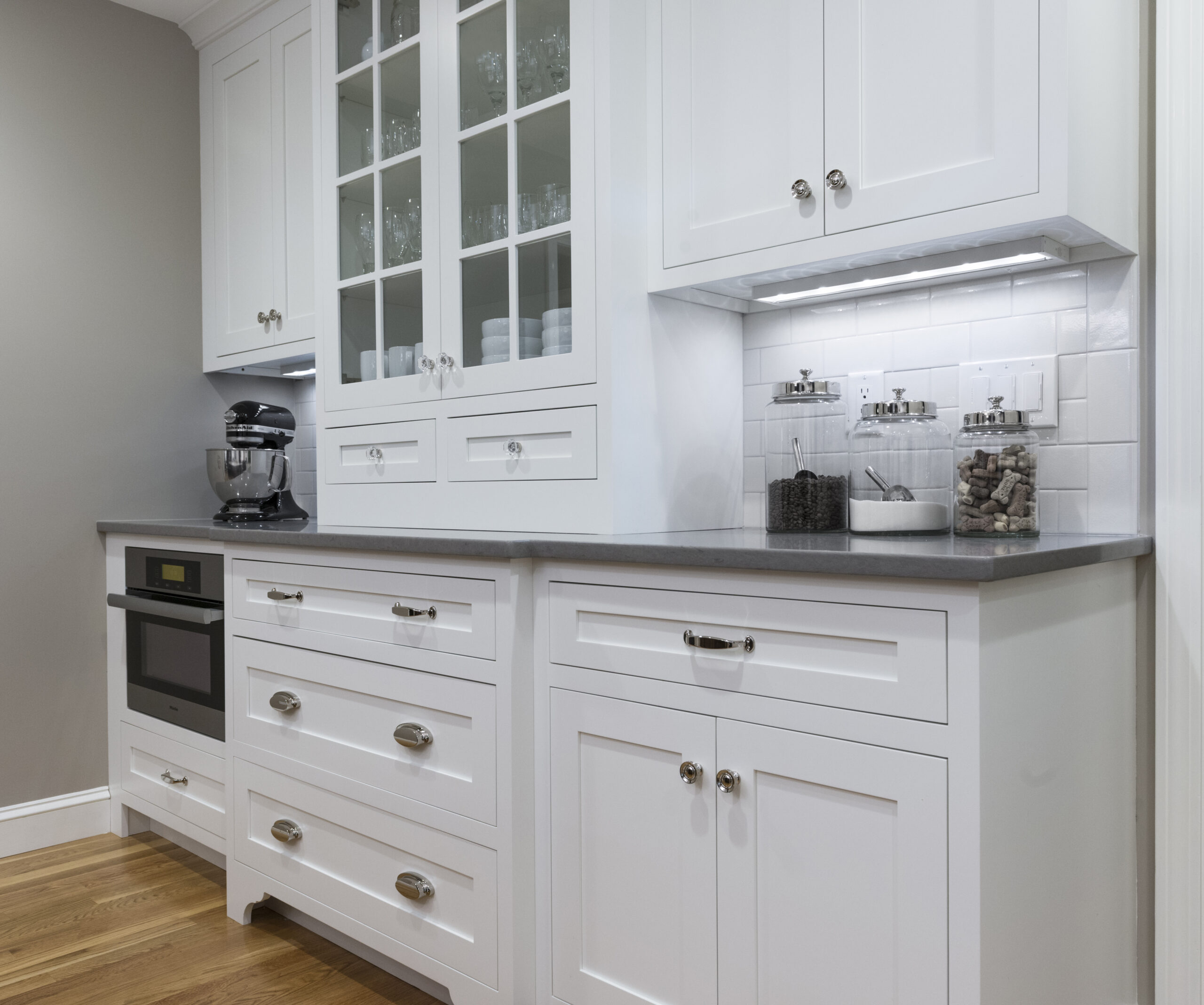 Modern kitchen with white cabinets, glass-front upper doors, a gray countertop, built-in microwave, stand mixer, and jars on the counter—showcasing the touch of an expert Interior Designer Massachusetts against light walls and a wood floor.