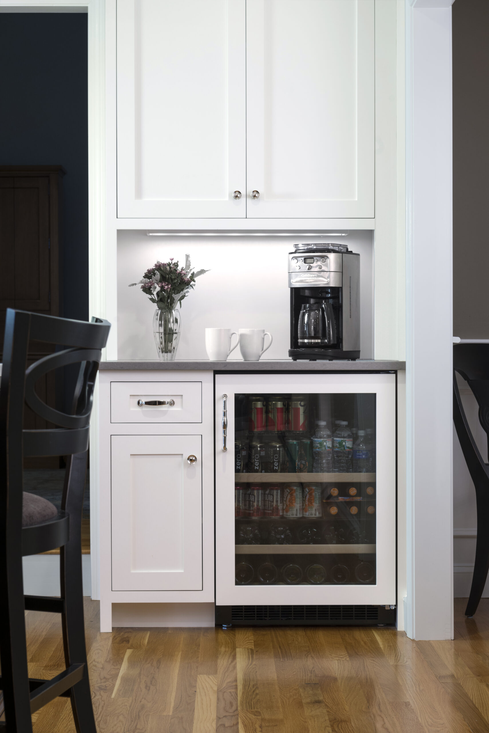 A small kitchen nook with white cabinets, a countertop holding a vase of flowers, two mugs, a coffee maker, and a mini fridge stocked with drinks underneath—perfectly styled for luxury interior design.