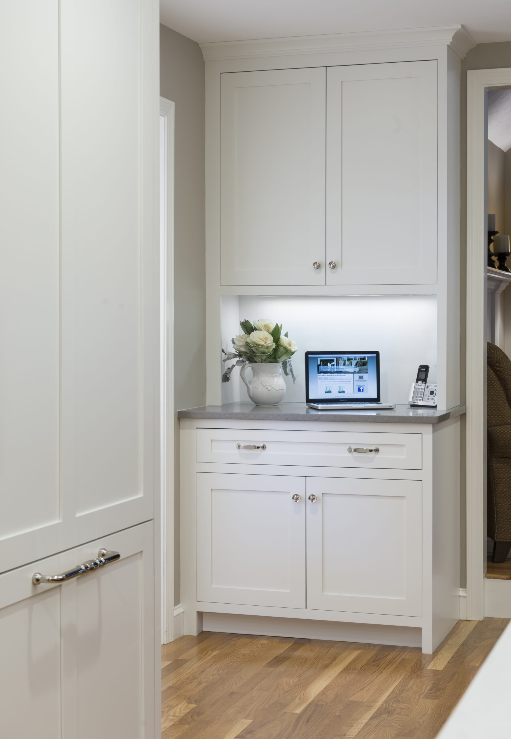 A built-in white kitchen cabinet with upper and lower doors, a vase of flowers, a laptop, and a cordless phone on a gray countertop showcases the elegance of luxury interior design against a light-colored wall and wood floor.
