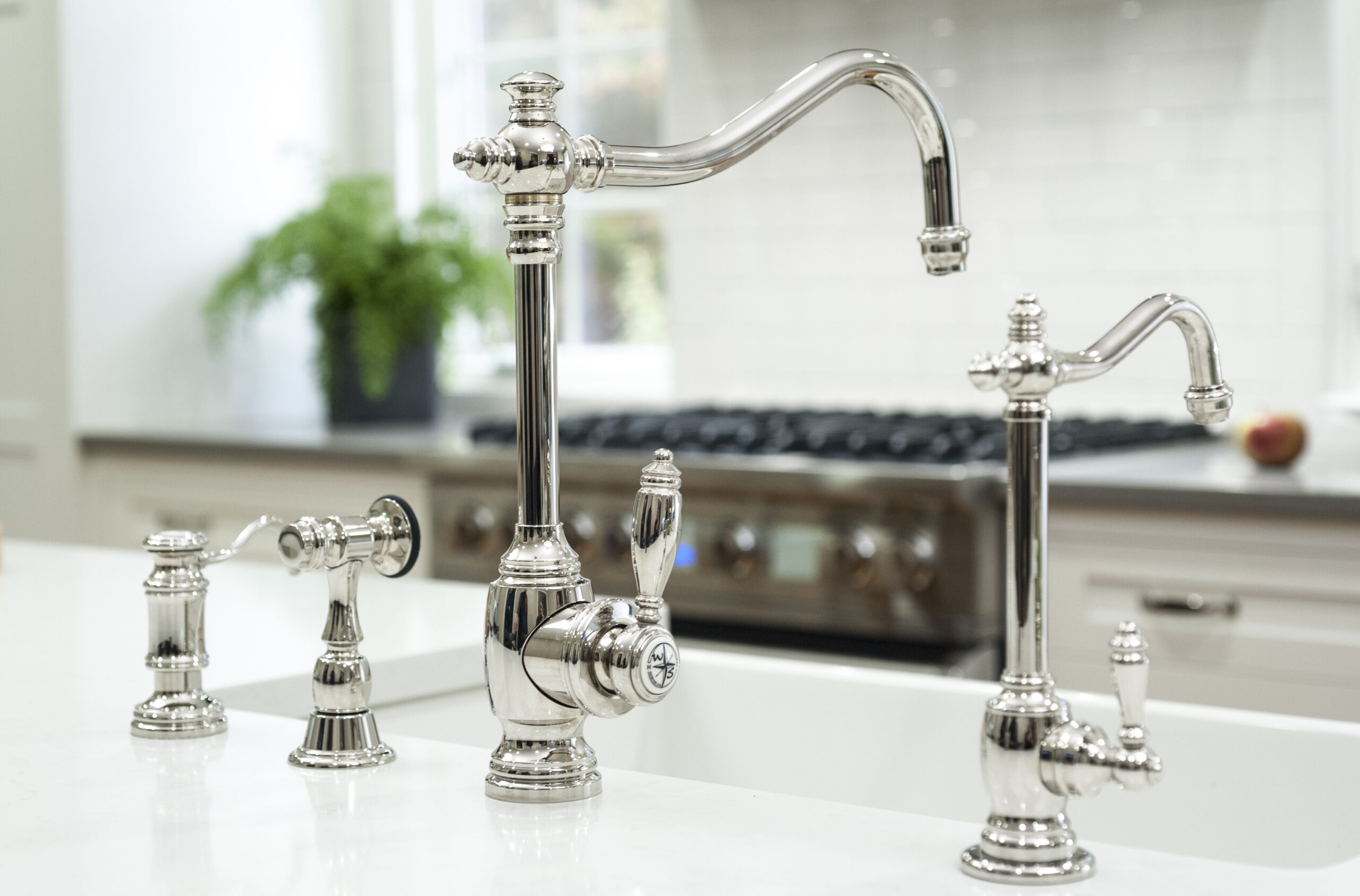 A close-up view of polished silver kitchen faucets and handles on a white countertop, reflecting the elegance of luxury interior design, with a stove and plant visible in the blurred background.