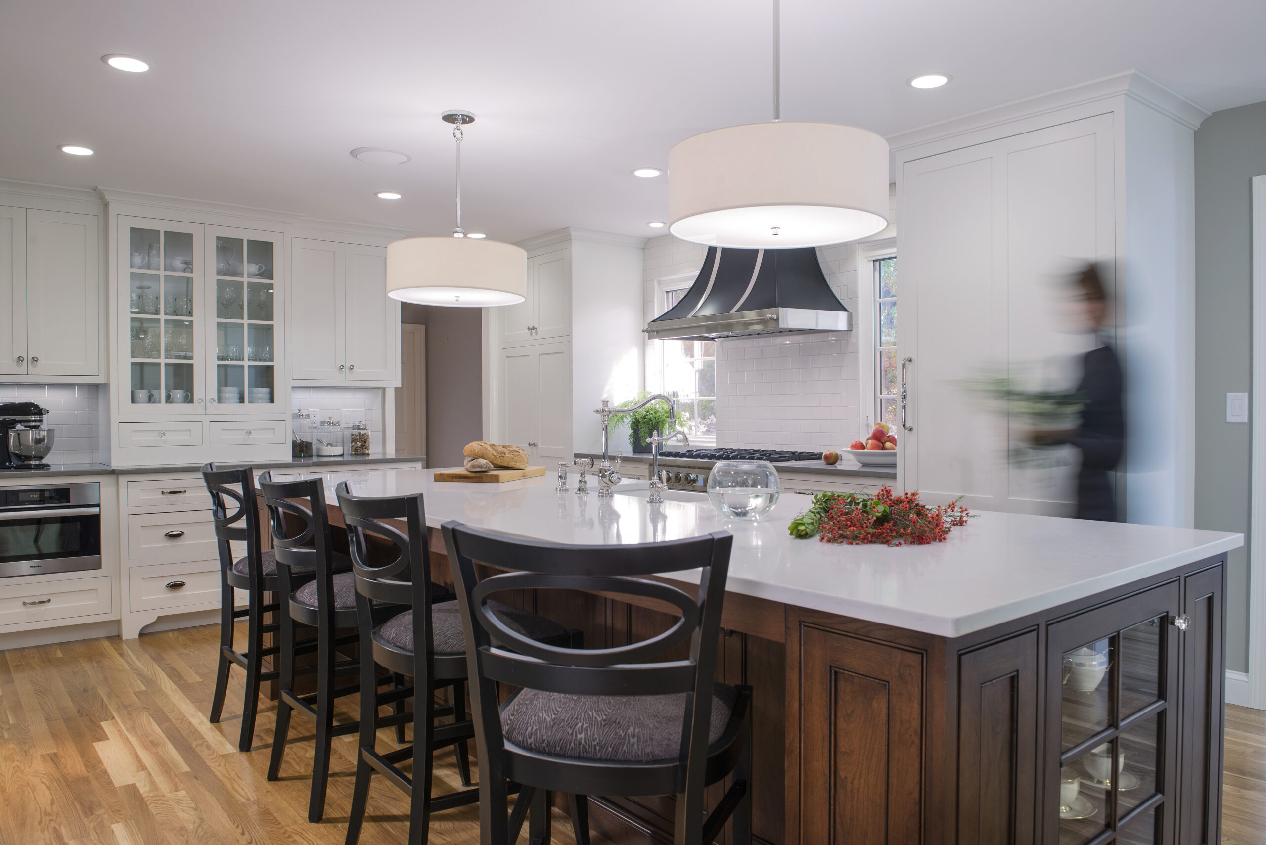 Modern kitchen with white cabinets, a large island with dark wood base, black chairs, pendant lights, and a blurred person walking near the stove holding greens—a stunning example of Luxury Interior Design by an Interior Designer Massachusetts.
