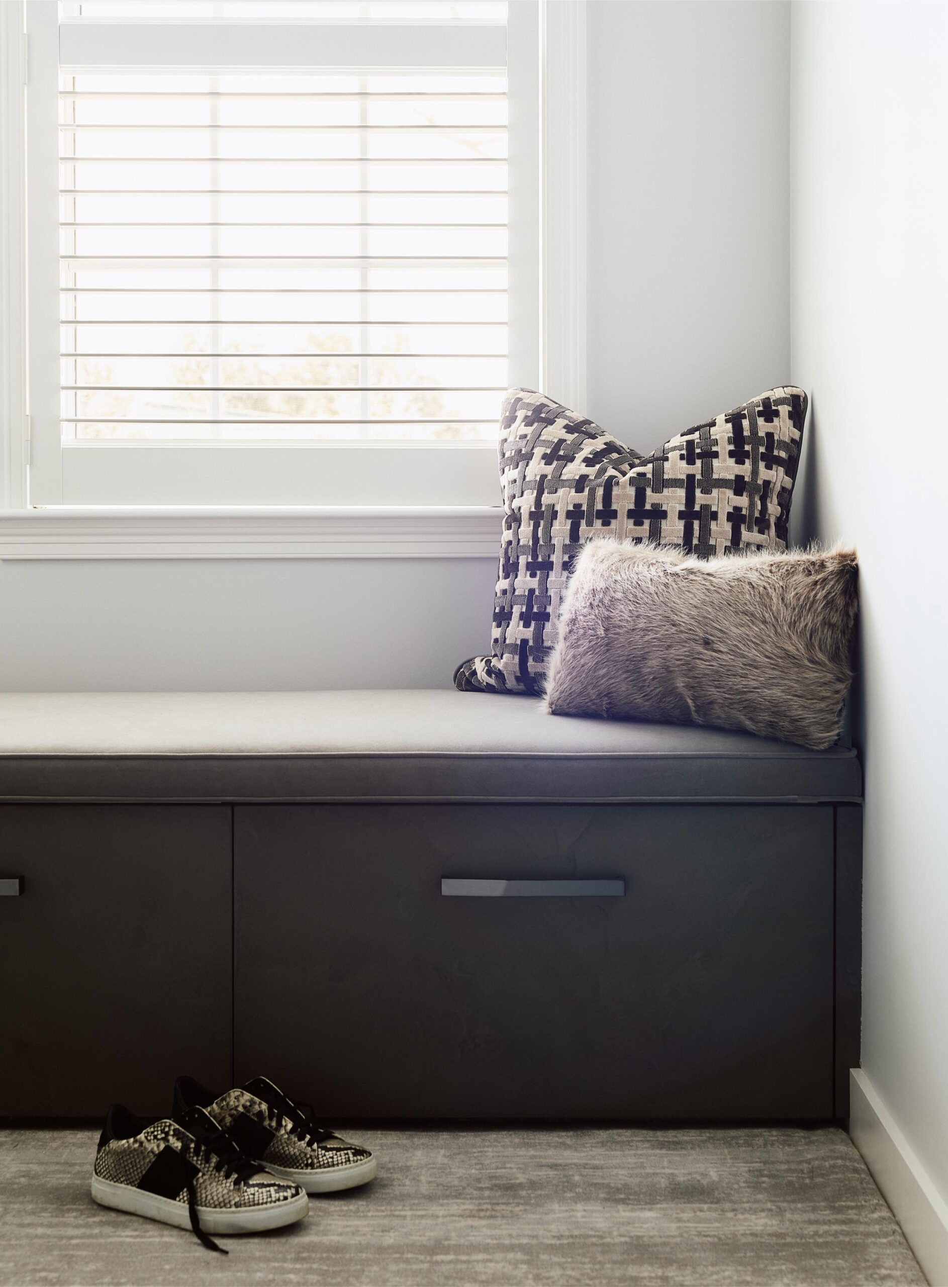 A pair of sneakers sits on the floor next to a built-in bench with two patterned cushions, showcasing thoughtful interior design beneath a window with white blinds.