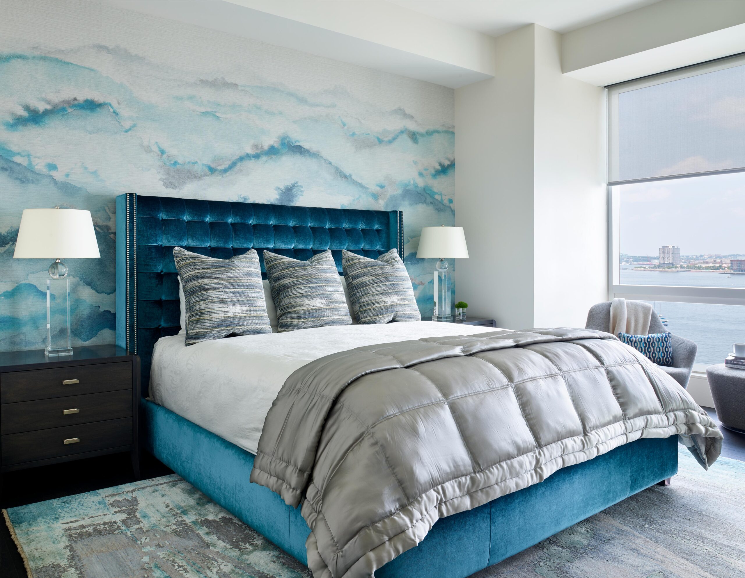 Modern bedroom with a blue upholstered bed, gray blanket, matching pillows, and stylish Interior Design touches. Features two bedside tables with lamps, a water-themed accent wall, and a large window showcasing city and water views.