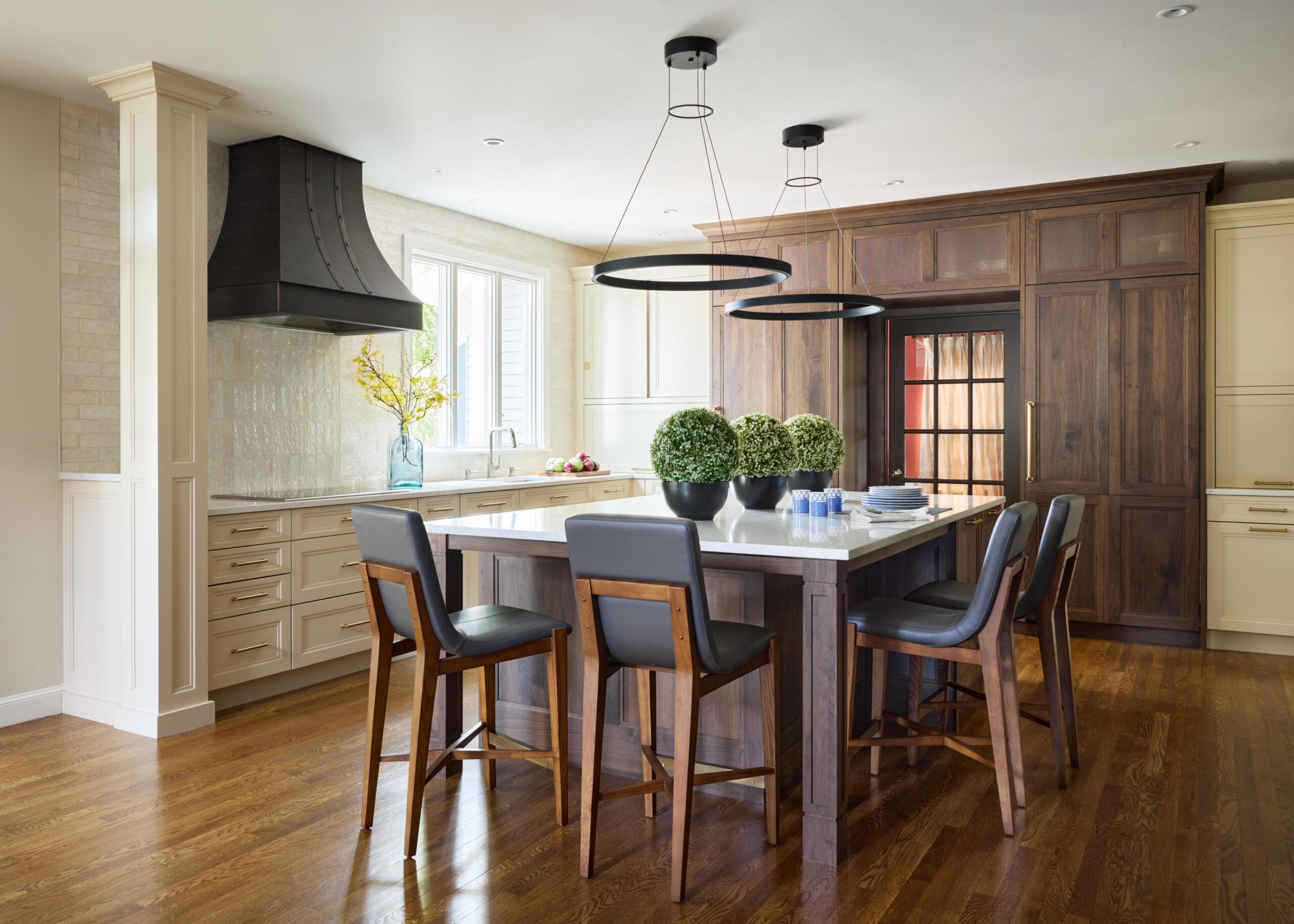 Modern kitchen with wood floors, a large island with four chairs, potted green plants, pendant lights, dark range hood, and built-in cabinetry around a spacious refrigerator—an inviting showcase of interior design.