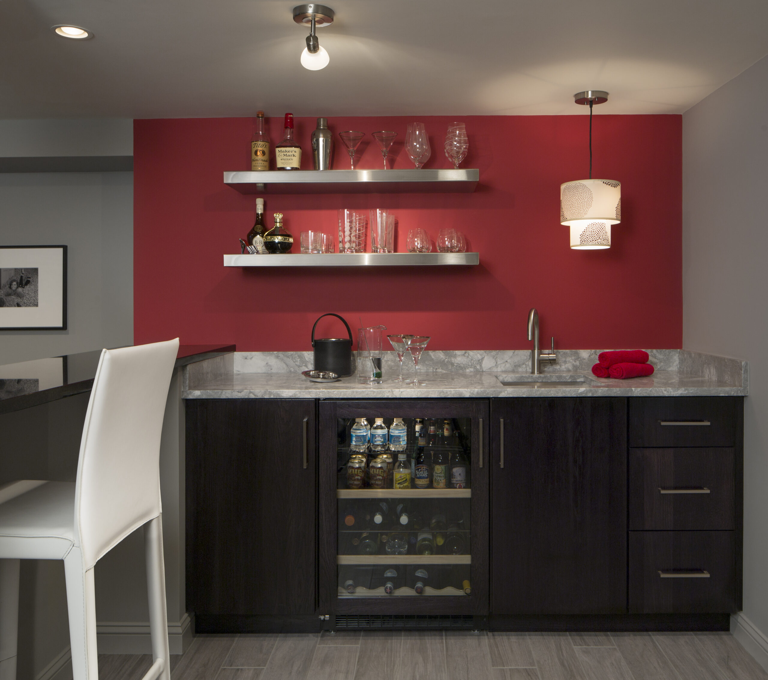 A modern home bar designed with luxury interior design flair features a red accent wall, glass shelves for bottles and glassware, marble countertop, sink, beverage fridge, and a white barstool—perfect inspiration from an interior designer in Massachusetts.