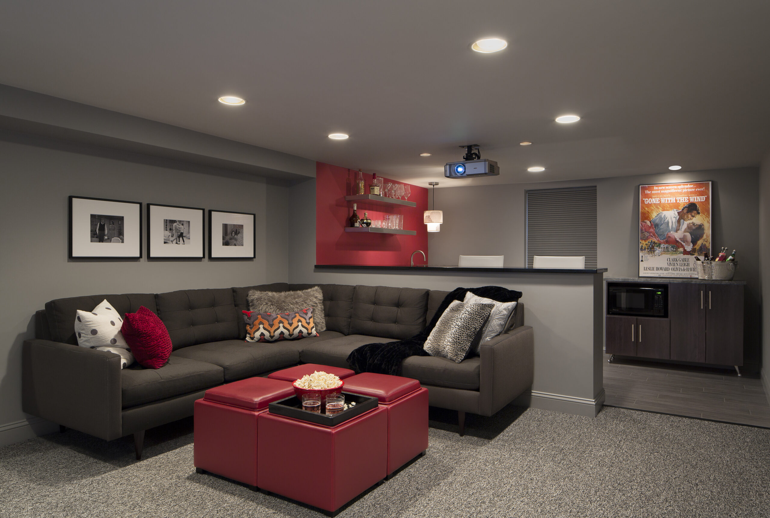 A modern home theater room with a gray sectional sofa, red ottomans, a projector, a snack bar area, and framed art on the wall—perfectly showcasing luxury interior design by an expert interior designer from Massachusetts.