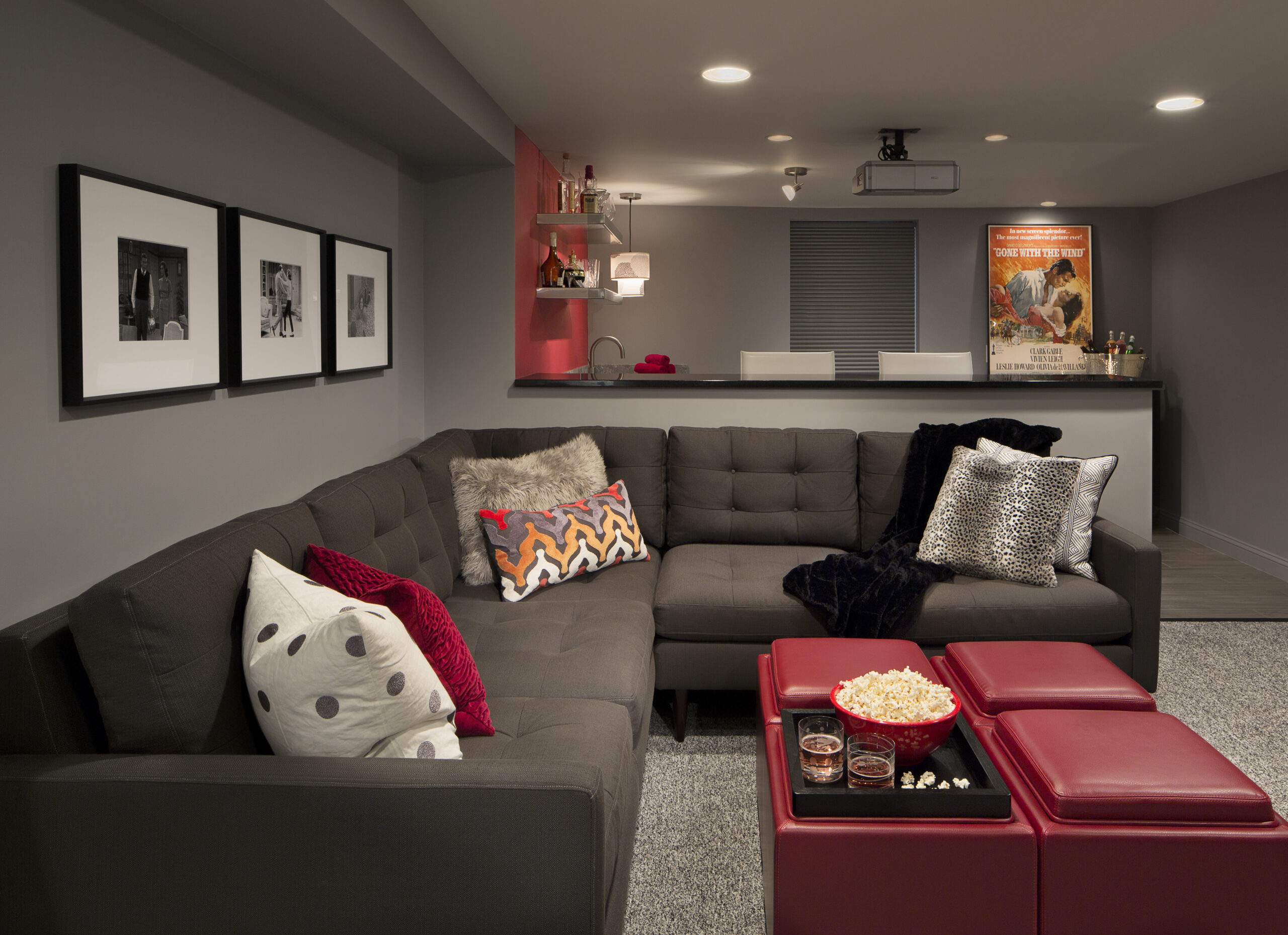 A modern basement home theater designed with luxury interior design touches—gray sectional sofa, red ottomans, framed art on gray walls, a stylish bar area, and a bowl of popcorn—shows the work of an expert interior designer in Massachusetts.