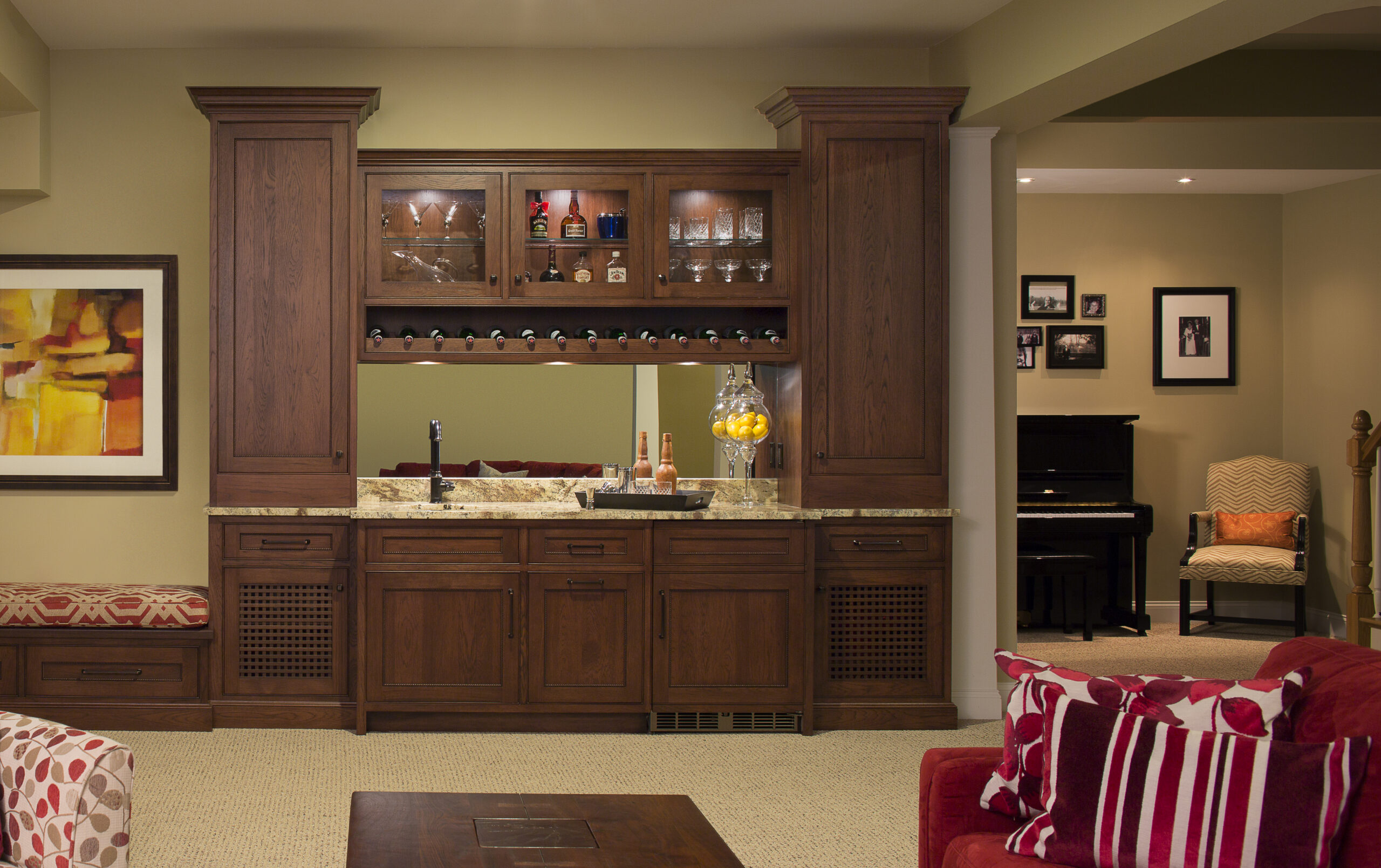 A home bar with dark wood cabinets, glassware, and bottles on display, set against a beige wall with a mirror—an inviting touch of luxury interior design near a living area with a piano and framed photos.