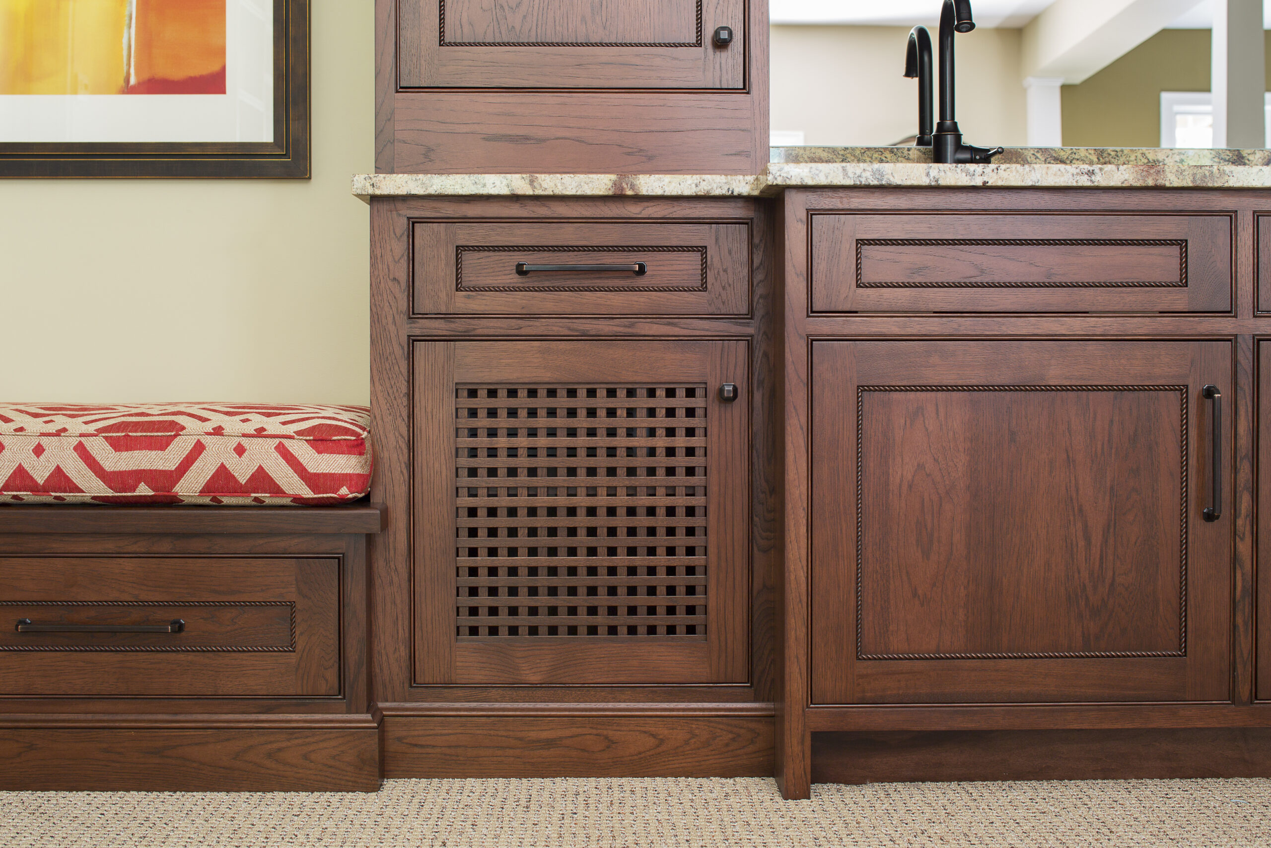 Wooden cabinetry with dark handles, a vented panel, and a beige granite countertop offers a touch of luxury interior design; a red patterned bench is partly visible on the left side.