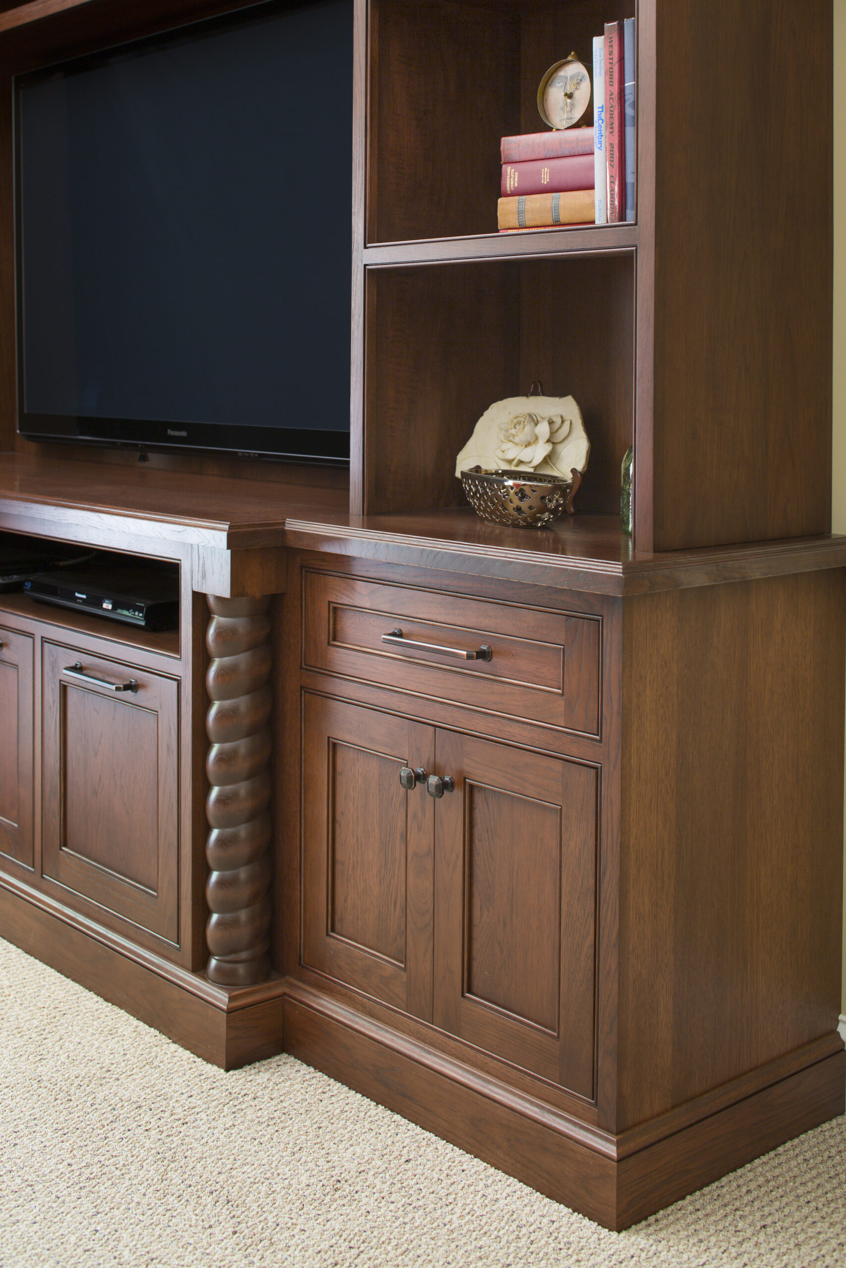 Wooden entertainment center with shelves, books, a decorative basket with a ceramic flower, a flat-screen TV, and cabinets on a beige carpeted floor—perfect for those seeking luxury interior design by an experienced Interior Designer Massachusetts.