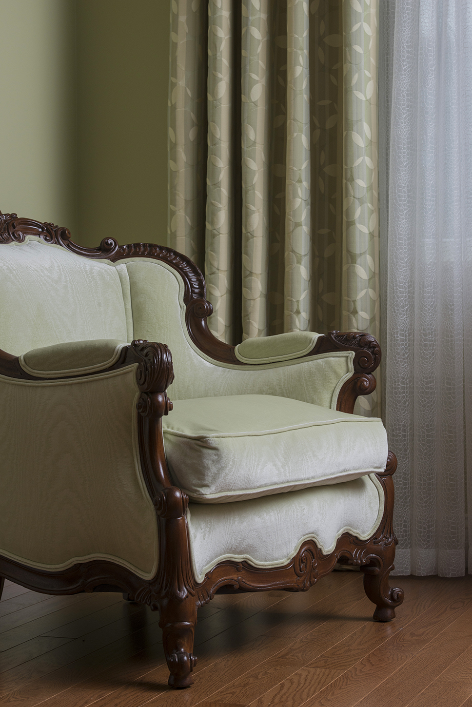 Ornate wooden armchair with light green upholstery beside patterned curtains and sheer white drapes on a wooden floor—an inviting touch of luxury interior design.