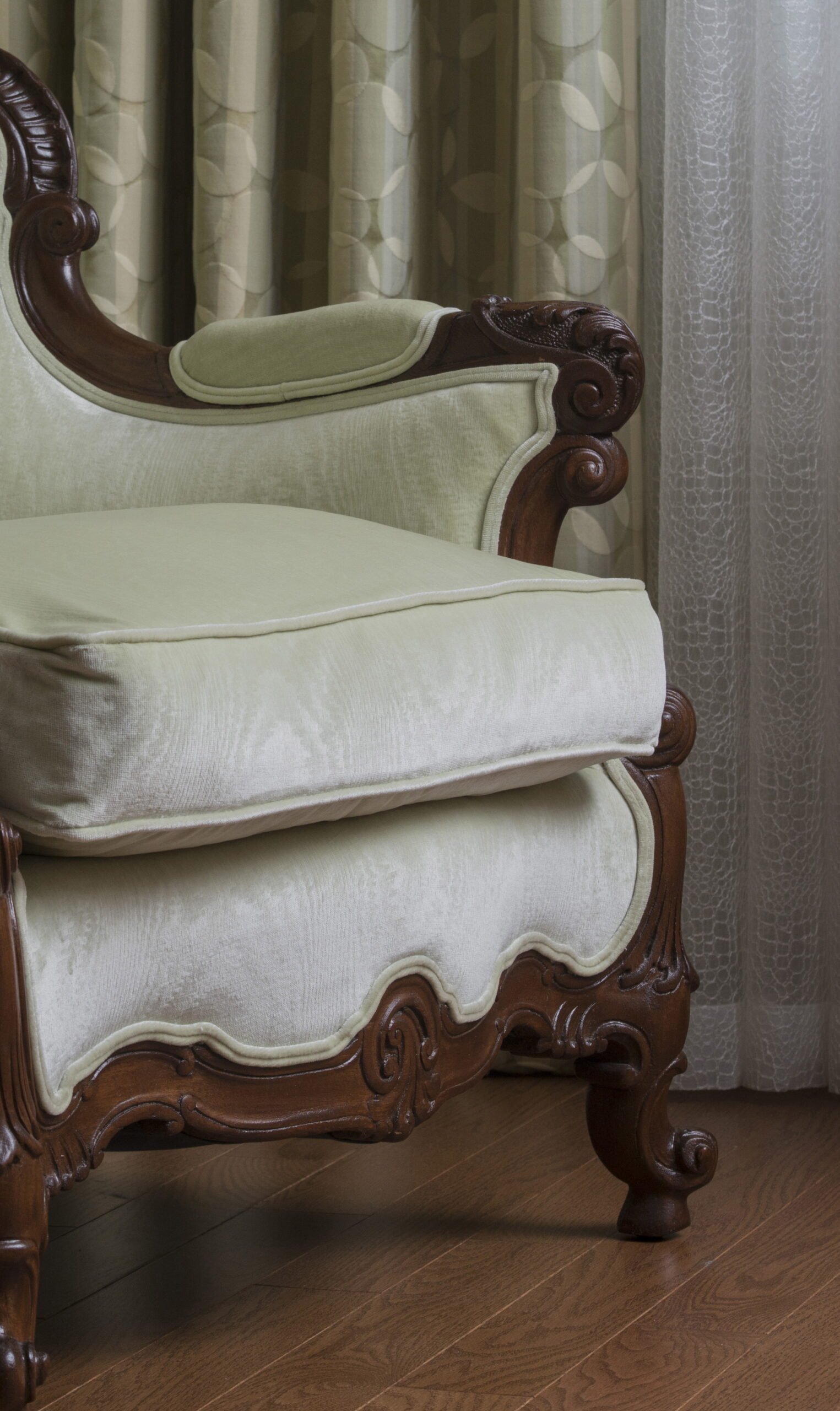Ornate wooden armchair with light green upholstery, placed on a wooden floor next to patterned curtains and sheer drapes—perfect for a Luxury Interior Design curated by an expert Interior Designer Massachusetts.