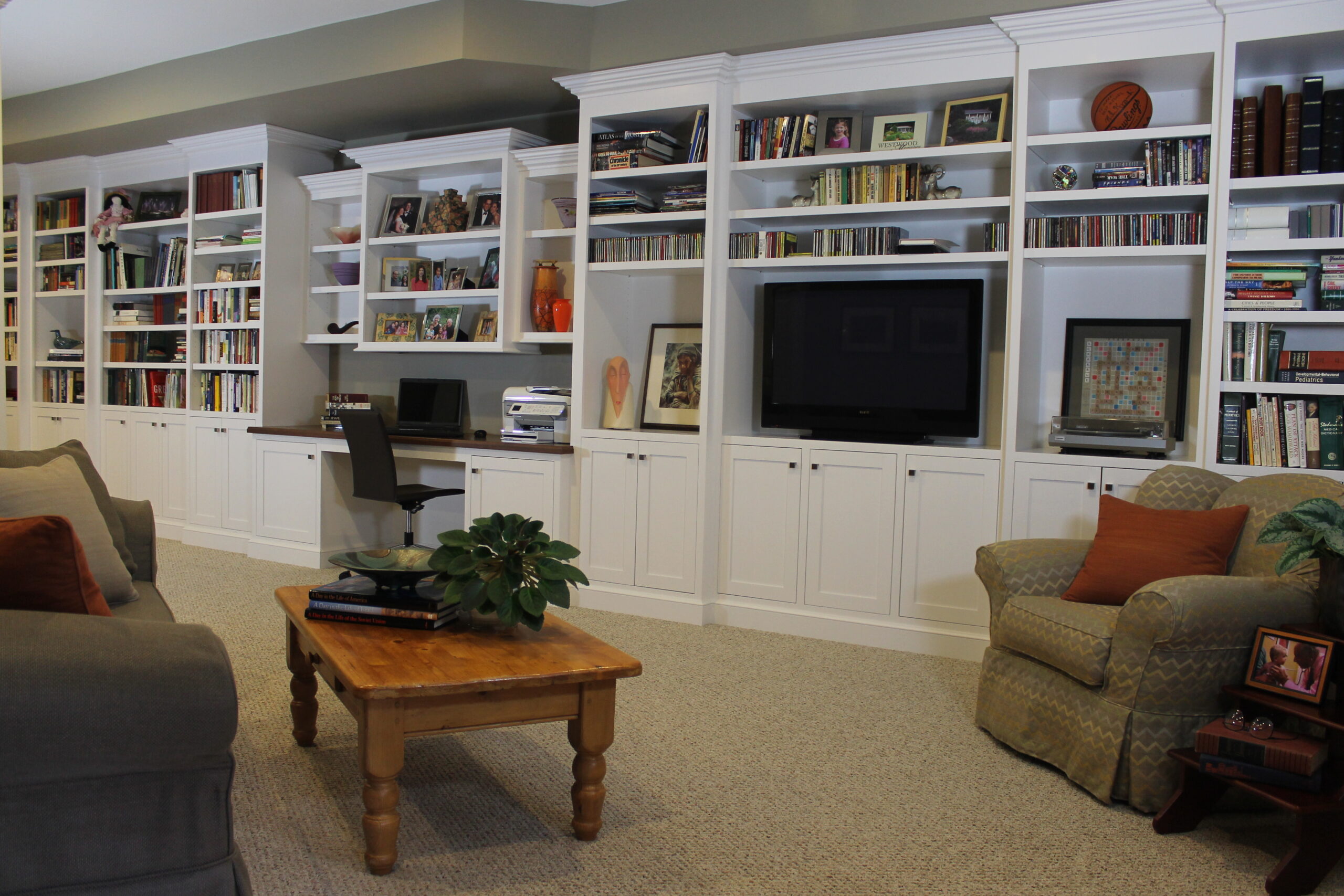 A living room curated by an Interior Designer Massachusetts features built-in white shelves, a TV, books, and decor. A desk, armchair, sofas, and a wooden coffee table with a plant create luxury interior design on carpeted flooring.