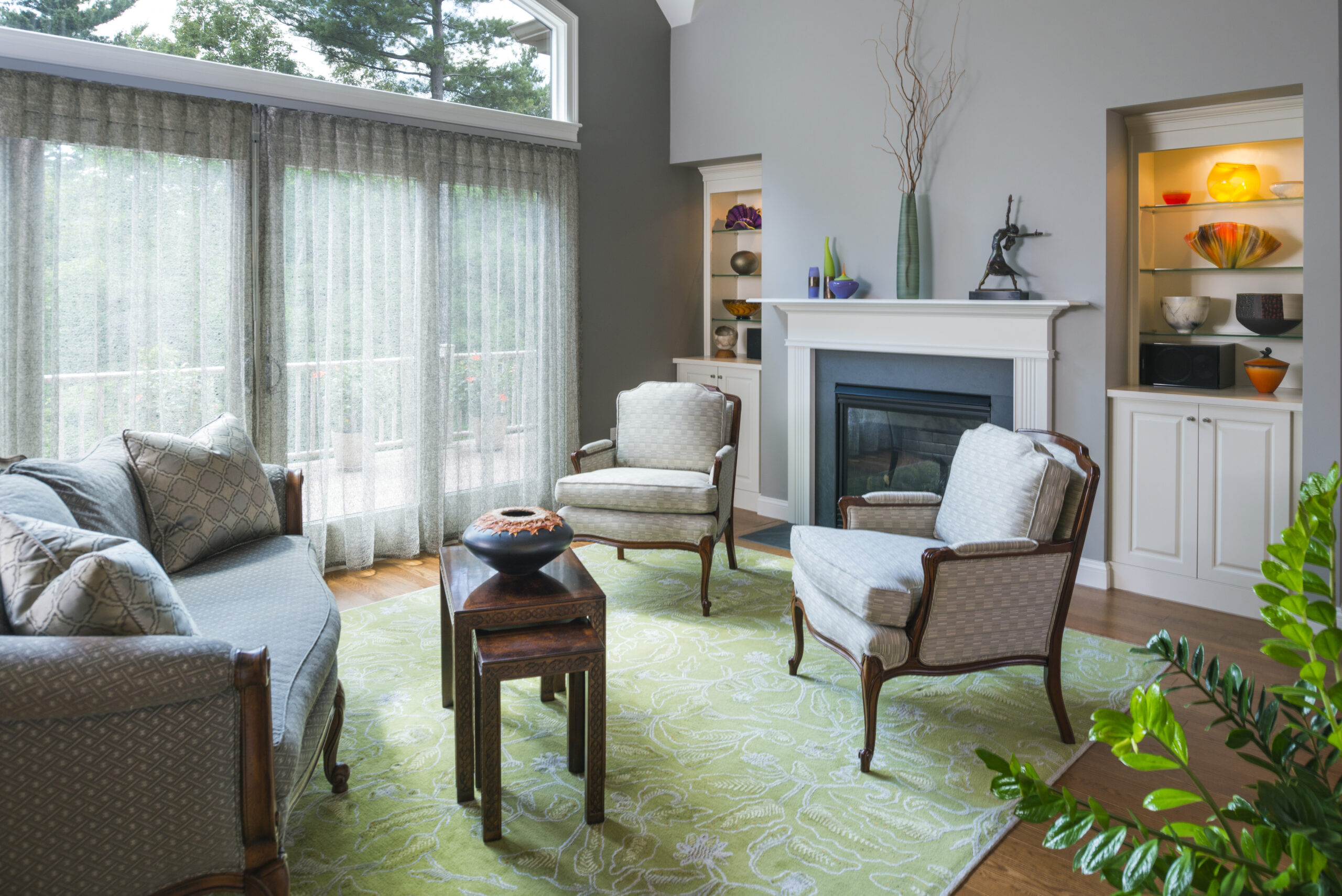 A bright living room with a green rug, sofa, three armchairs, fireplace, built-in shelves with decorative items, and large windows with sheer curtains showcases luxury interior design by an expert interior designer in Massachusetts.